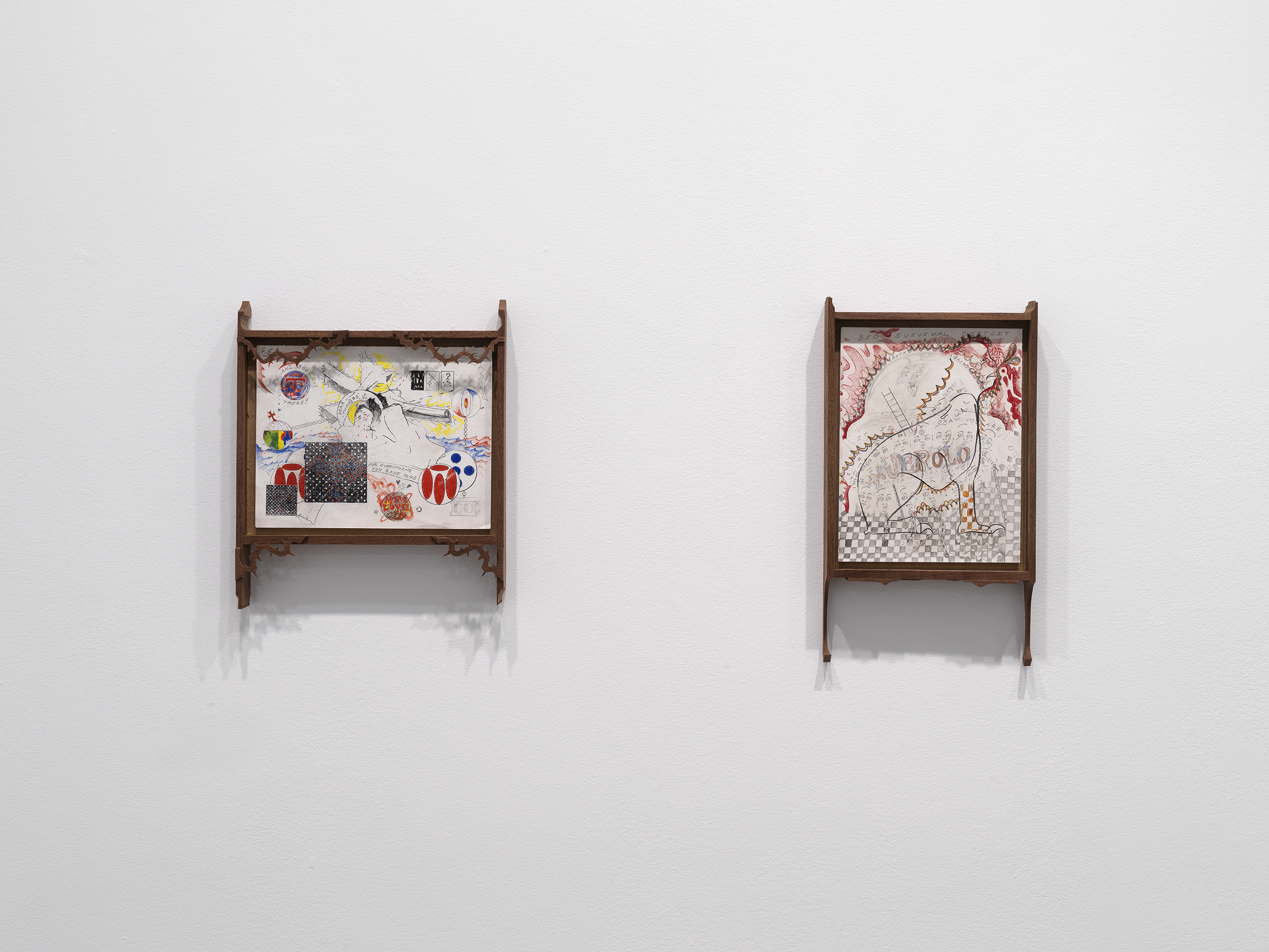 On a white wall hang two drawings in wooden frames. The left drawing is horizontal and has colorful abstract forms scattered across the paper, including forms that resemble globes, playing cards, and waves. The wooden frame is adorned with thorn motifs in each corner. The right vertical drawing depicts a four-legged animal-like form with red flame-like designs in the background and a gray and white checkerboard print at the bottom.