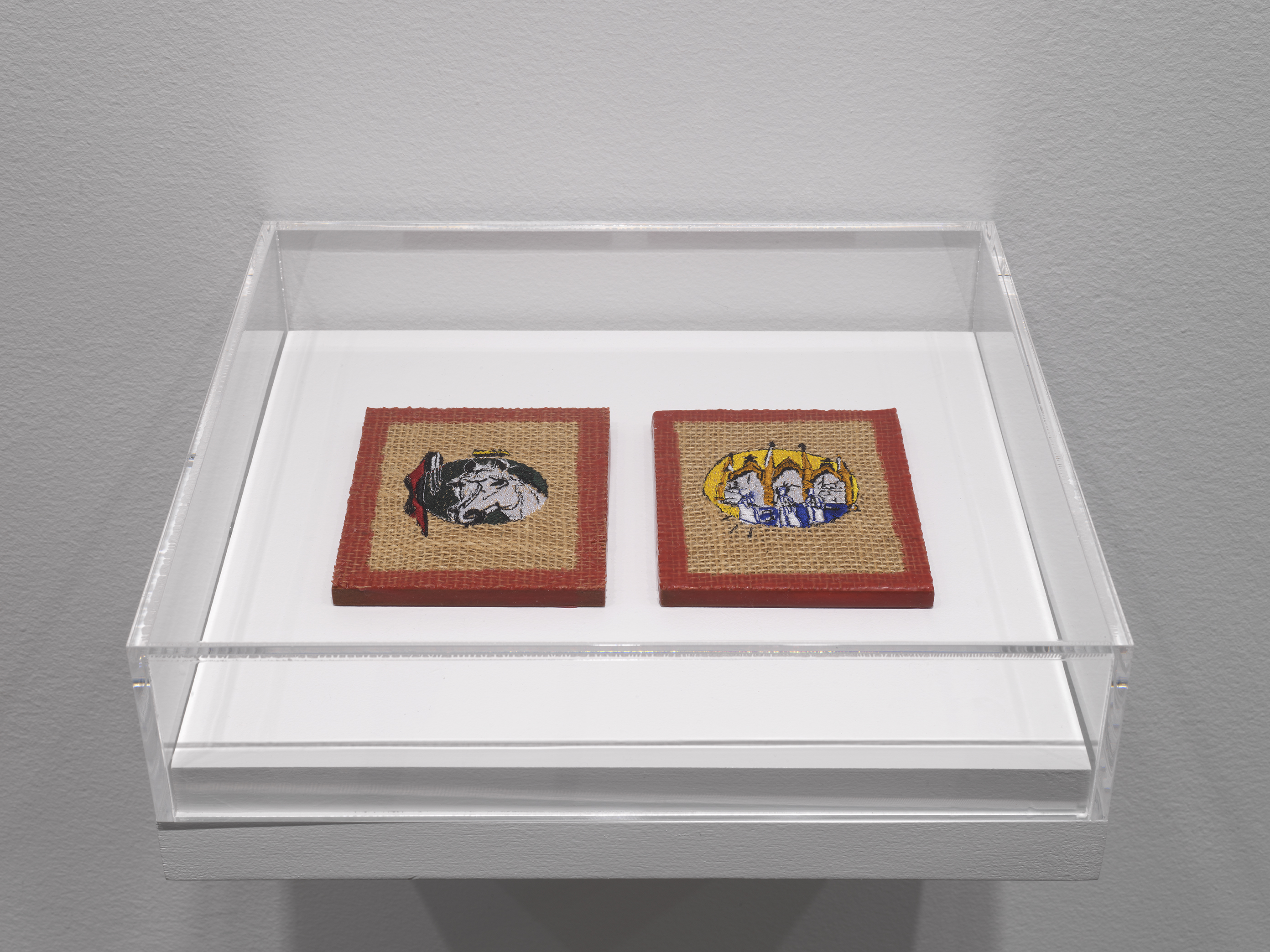 A plexiglass case hangs off of a white wall. Inside the case, two burlap-like square artworks are adorned with embroidered details. The left shows the decorated head of a horse, while the left is more ambiguous but has a similar medieval flavor. Each square has a border of red wax around the piece.