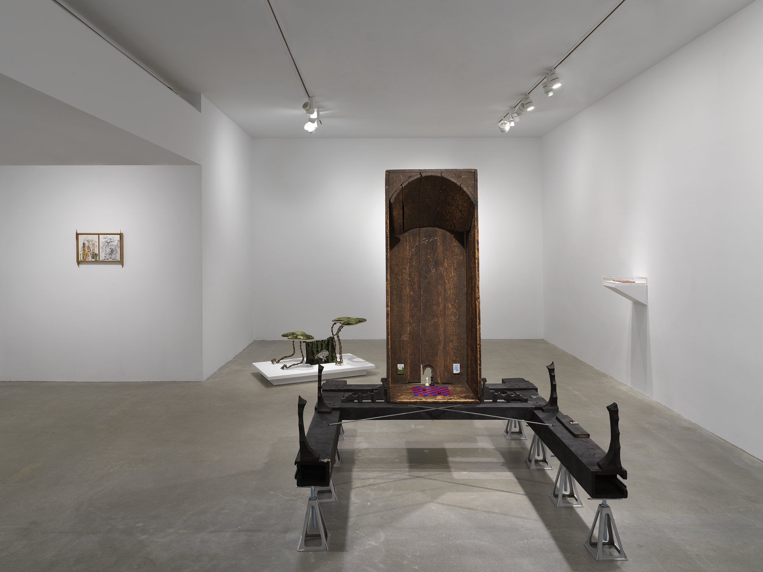 In the center of the room, a vertical wooden bathtub sits on horizontal wooden beams, which are held up by eight metal supports. Inside the bathtub are small painted scenes and a red and blue checkerboard pattern on the base. On the left wall, a small diptych drawing in a wooden frame hangs. In the background behind the wooden bathtub, a small creature-like sculpture made of metal and fabric sits on a low pedestal. On the right, a white shelf hangs off the wall with a plexiglass case on top.