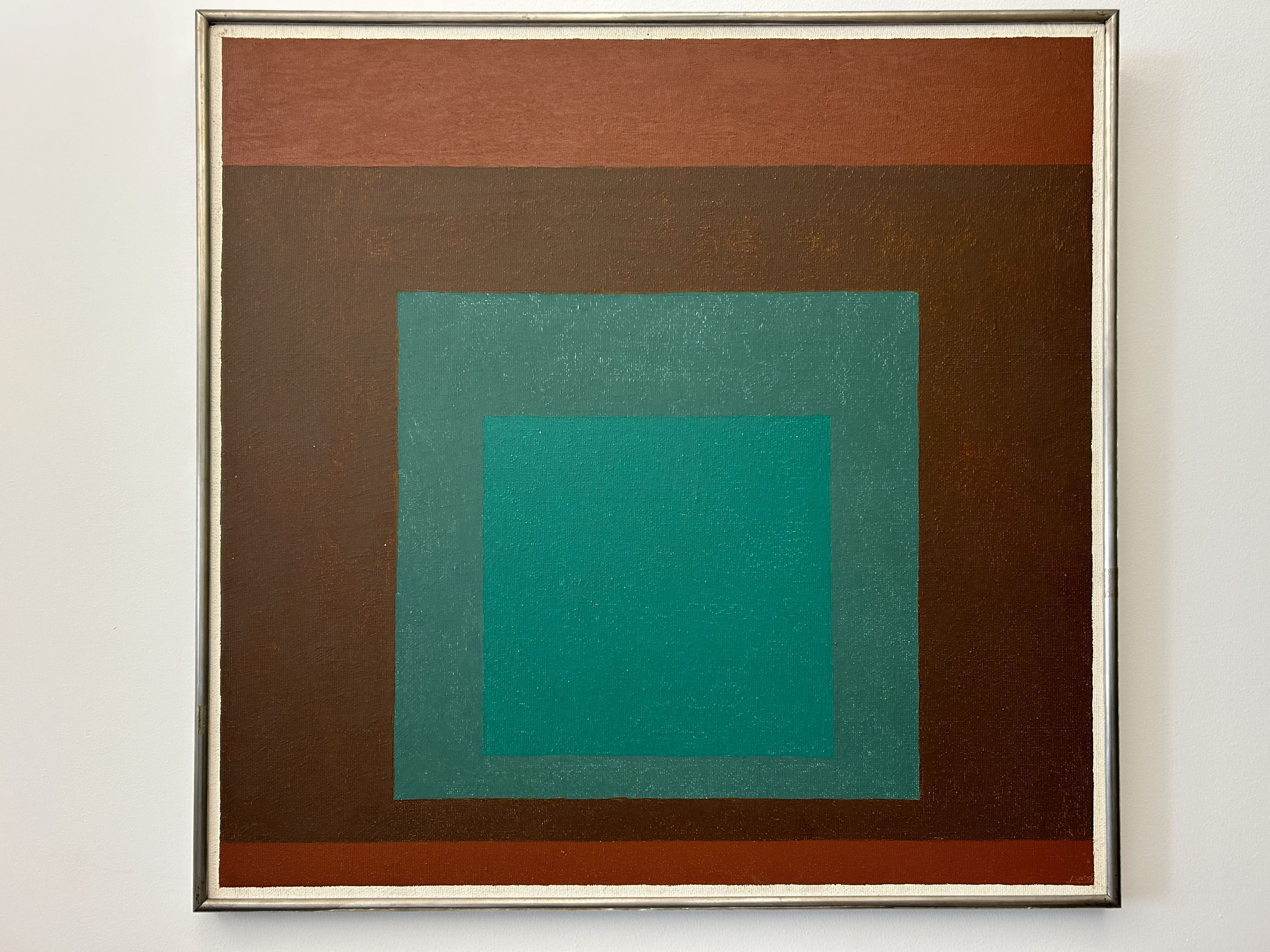 Square painting with a silver frame on a white wall of a brown background with a smaller turquoise square in the center and a lighter brown stripe across the top.