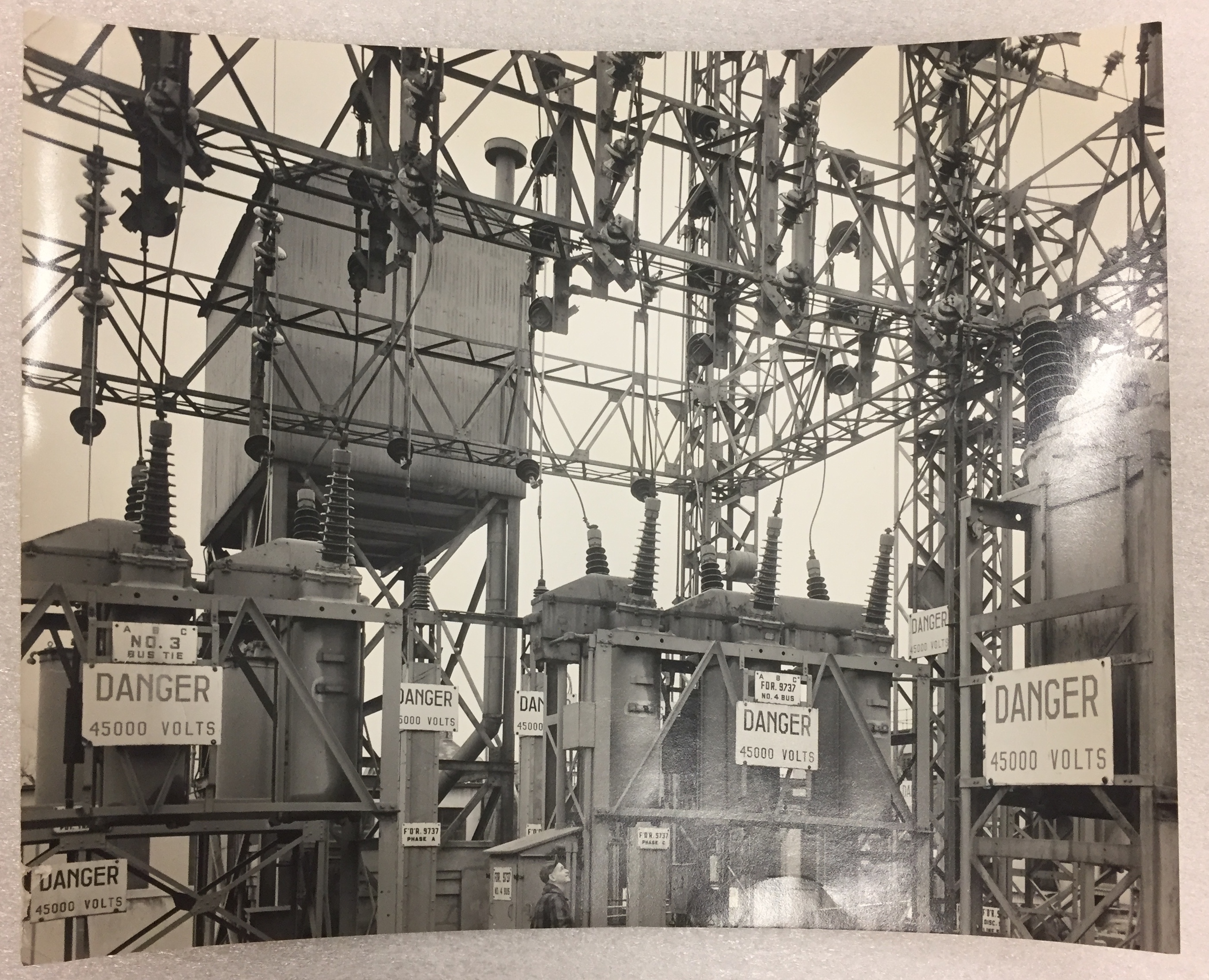 Black and white photograph of a collection of transformers with nine danger signs hung on various heights.