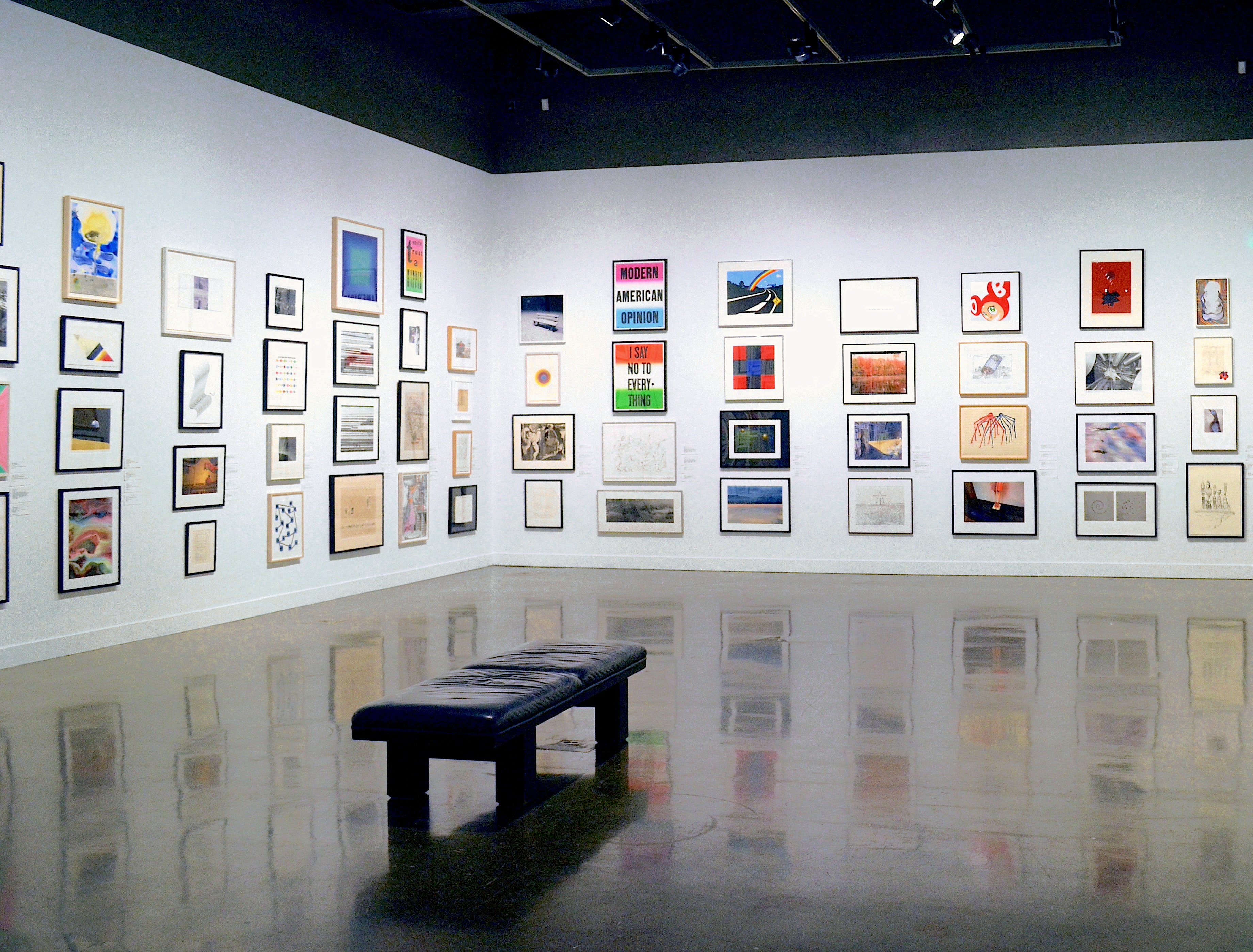 Installation view with framed artworks lining the walls floor to ceiling and a leather bench in the center of the gallery.