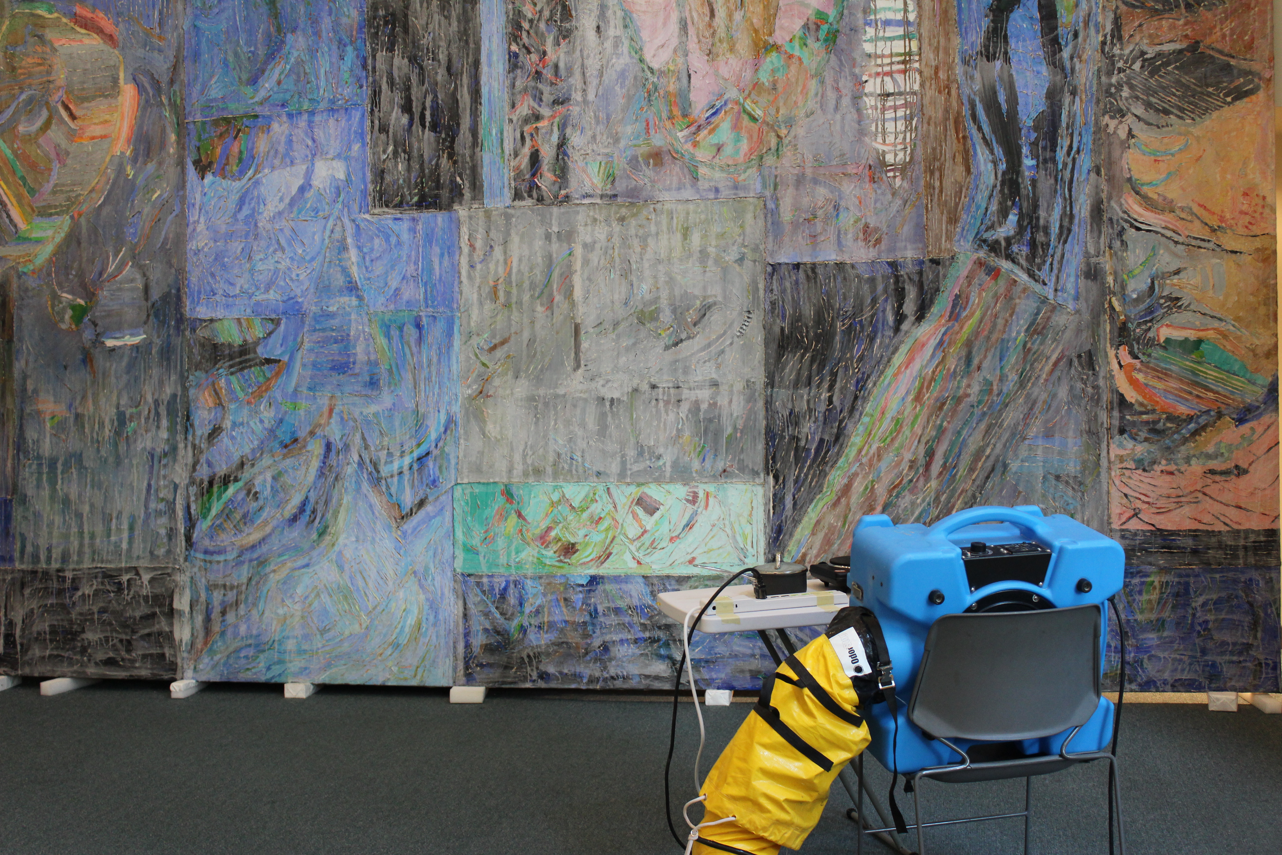 Large oil on canvas painting leaning against a white wall undergoes restoration. A small blue machine with a bright yellow vent tube is on a table in front of the painting on the right lower side of the image.