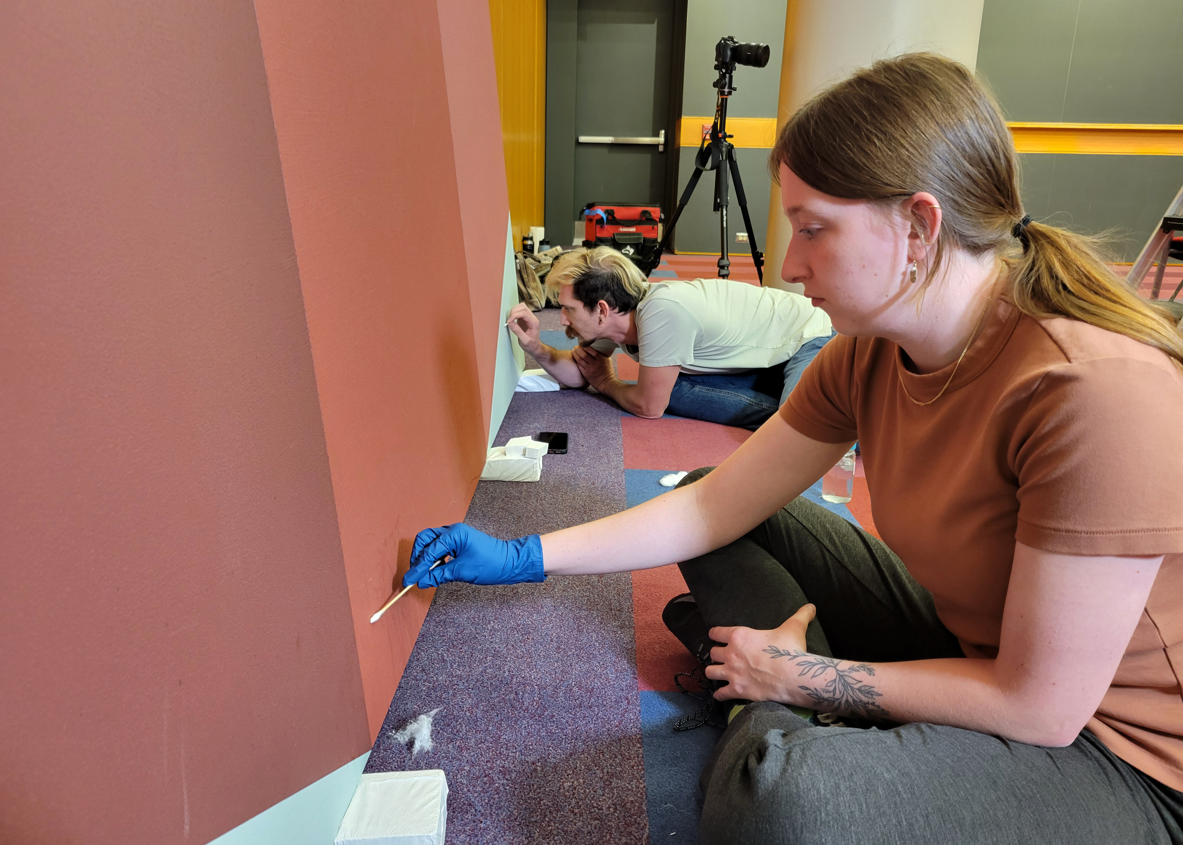 Image of two conservators sitting on the floor wearing blue gloves working on repairing a large scale red painting.