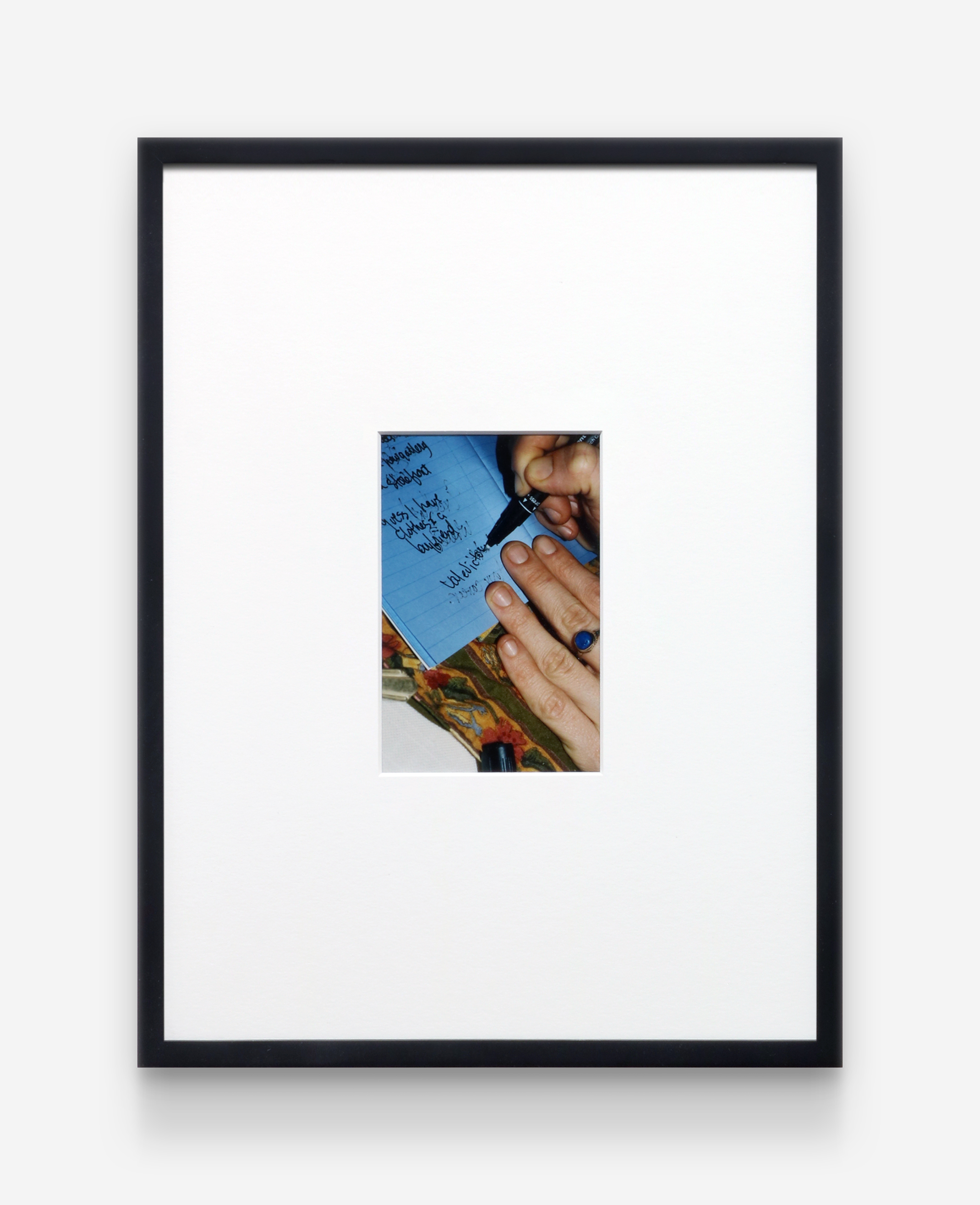 Print in a black frame with a thick white matte. The central photograph is of hands with a royal blue stone silver ring writing using a black ink pen on white lined paper.
