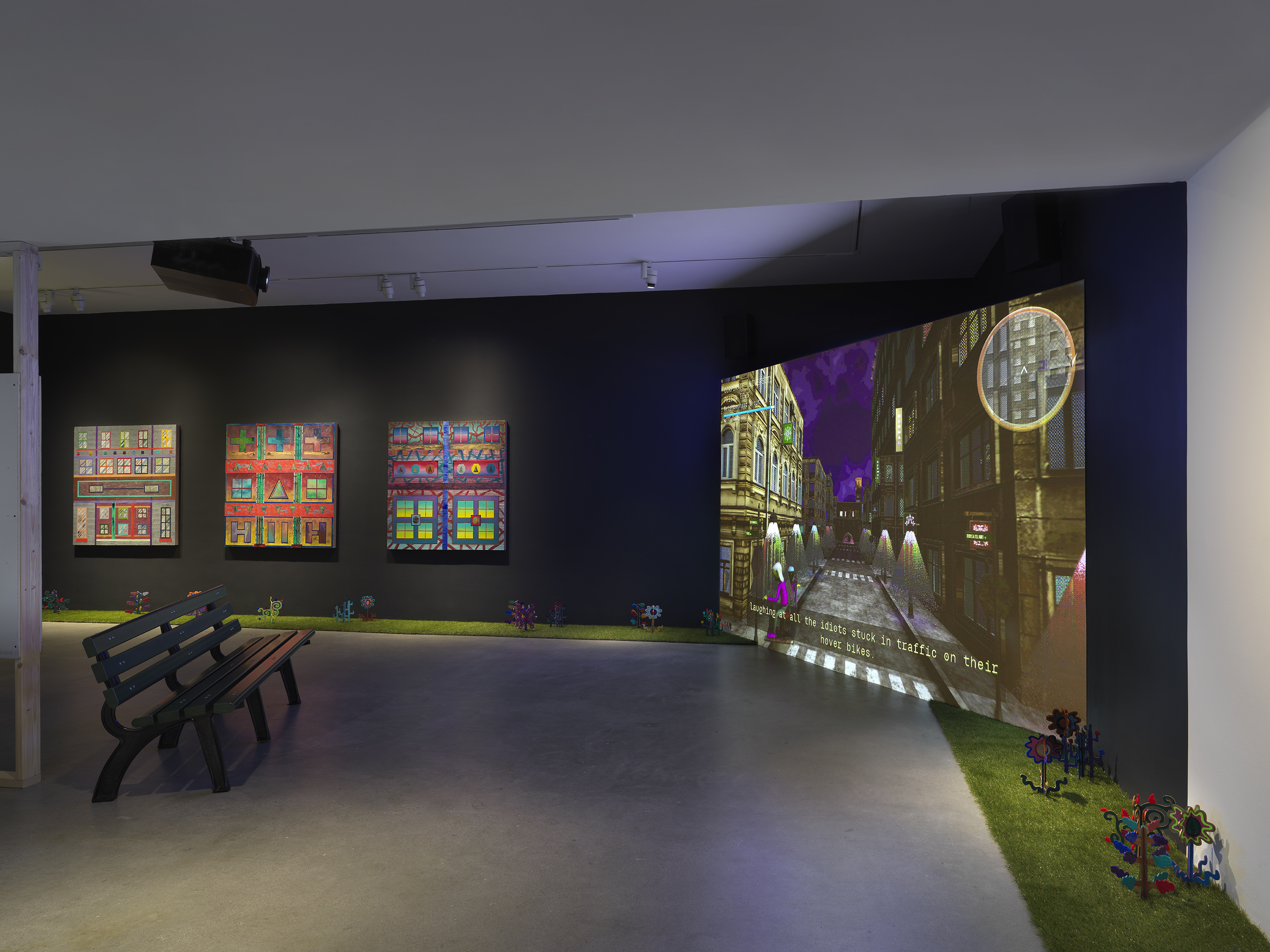 A dark gallery space with black walls is illuminated by a large screen. Digitally  rendered footage of a dark city street is projected onto the large screen in the right  corner of the room. A park bench is placed on the left side of the image facing the  screen. In the background, three square paintings depicting bright, geometric forms  hang in a row. Astroturf lines the walls of the room with painted wooden plants emerging  from it. 