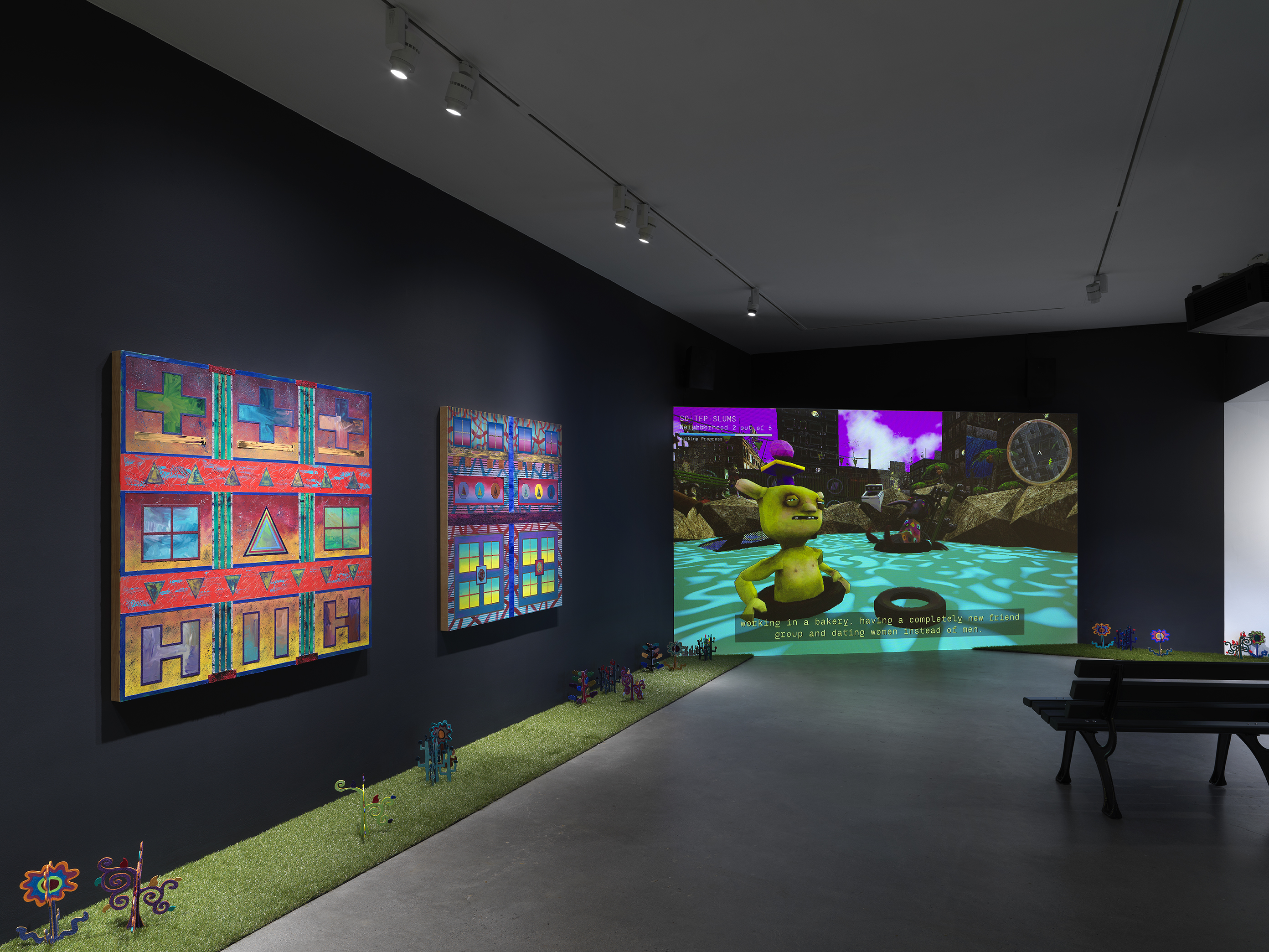 A dark, black-walled gallery space with artworks on display. On the left wall, two  paintings hang in a row depicting bright, geometric forms. In the back of the gallery, a  large screen illuminates the space displaying digitally rendered footage of a green  creature in water. A park bench faces the screen. A strip of astroturf lines the floor  around the gallery, with painted wooden flowers emerging from it.