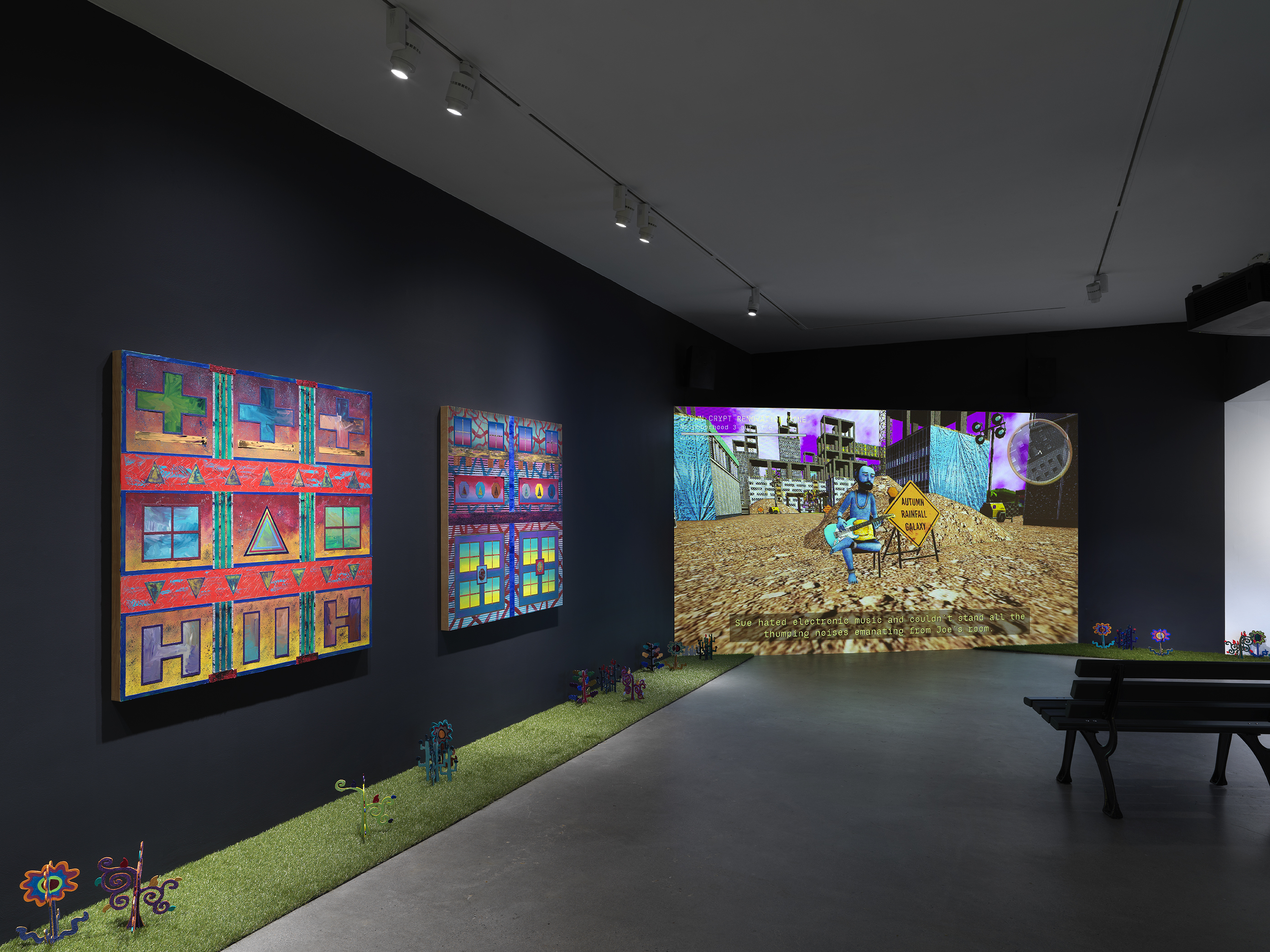 A dark, black-walled gallery space with artworks on display. On the left wall, two  paintings hang in a row depicting bright, geometric forms. In the back of the gallery, a  large screen illuminates the space displaying digitally rendered footage of a blue man  playing a guitar in the middle of a construction zone. A park bench faces the screen. A  strip of astroturf lines the floor around the gallery, with painted wooden flowers emerging  from it. 