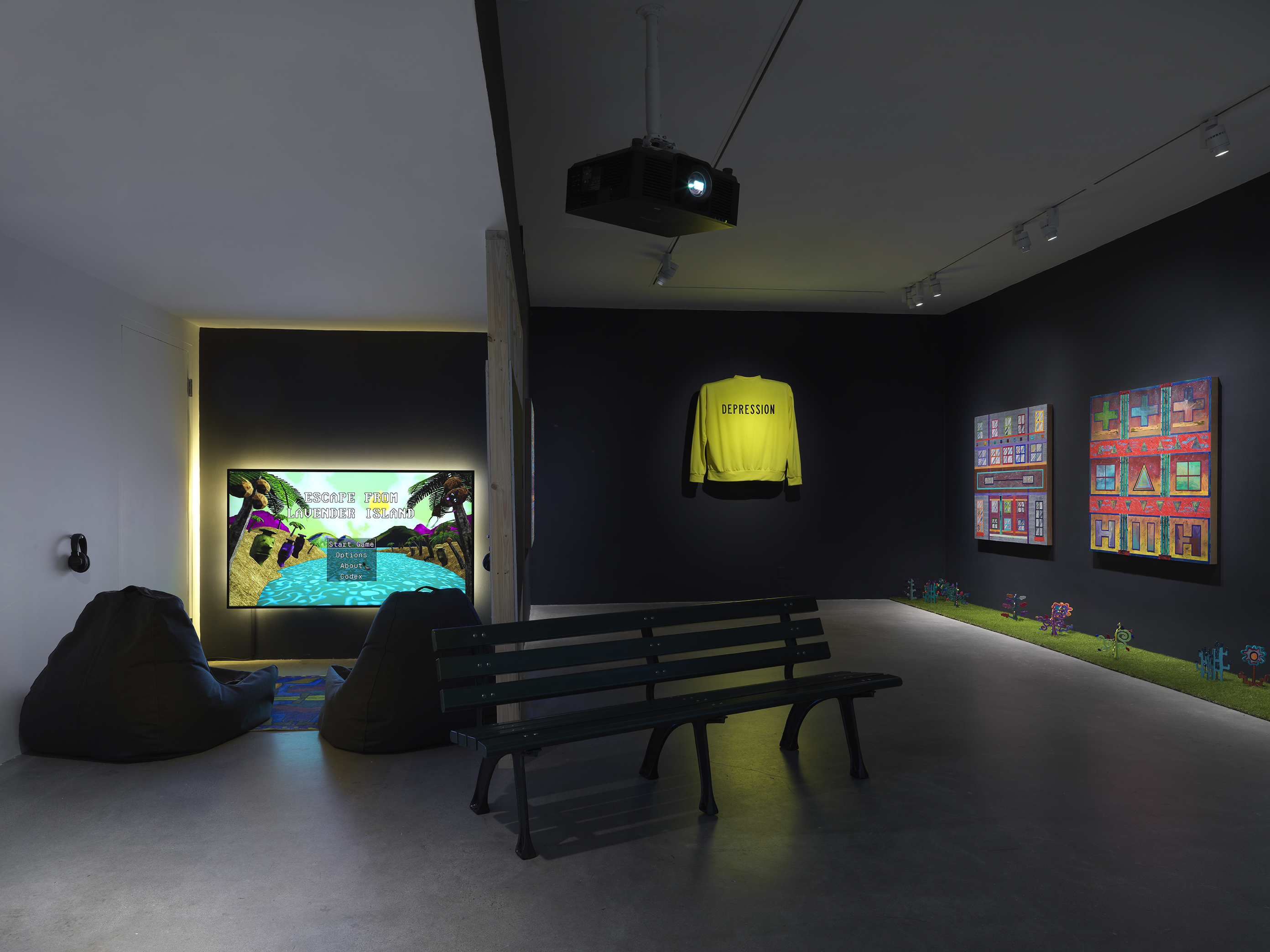 A dark gallery space with black walls is illuminated by a glowing TV on the back  wall with a video game menu displayed on the screen. In the center of the room, a  projector hangs from the ceiling above a park bench. On the back right wall, two square  paintings hang in a row. To the left of the paintings, a bright yellow sweater hangs elevated off the wall. 