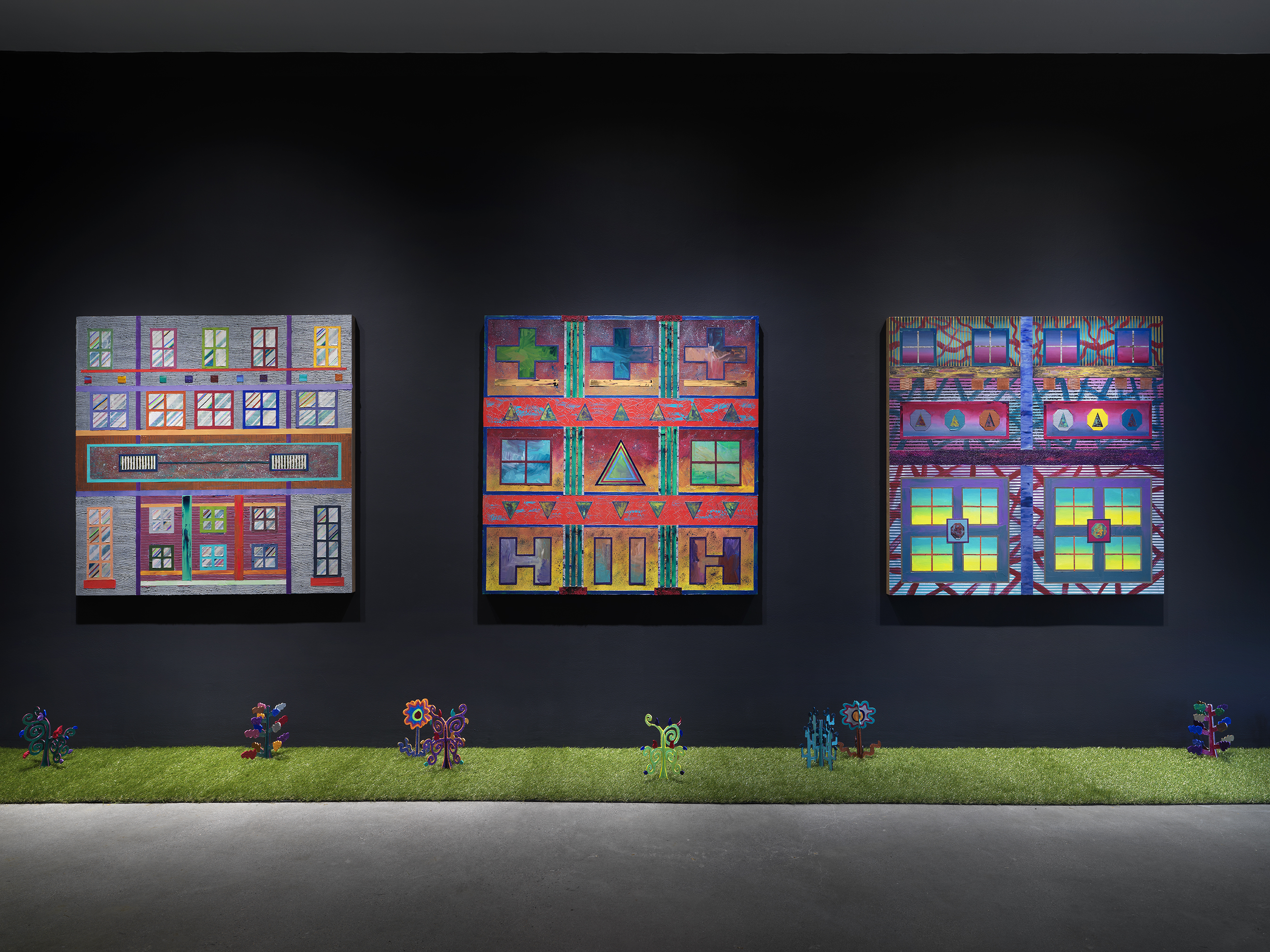 Three square paintings hang in a row on a black wall. The paintings depict  bright, geometric forms. A row of astroturf lines the floor against the wall, with small painted wooden plants emerging from it. 