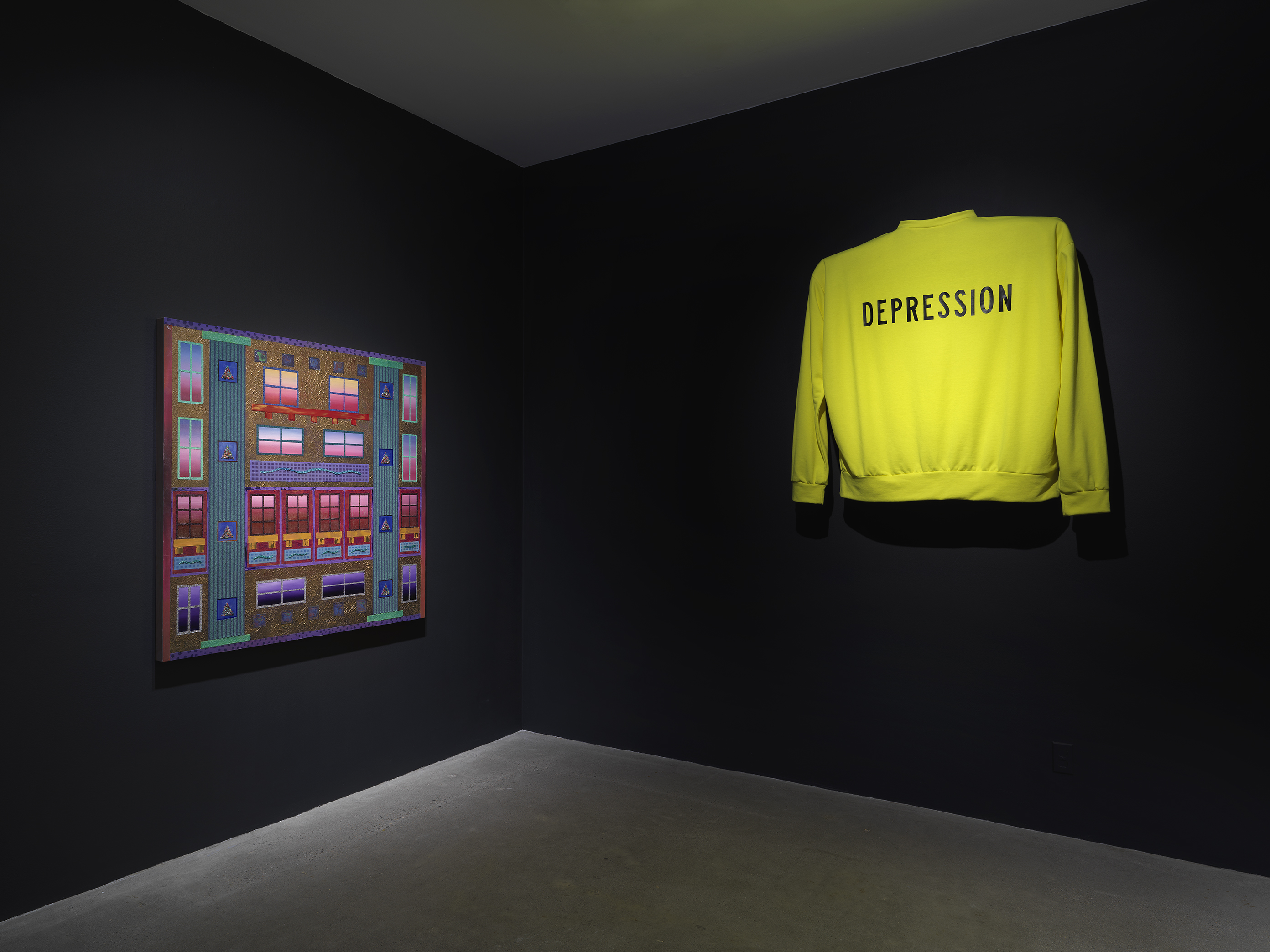The corner of a dark gallery space with black walls. On the left wall, a square  painting depicting bright, geometric, window-like forms is displayed. On the right wall, a  bright neon yellow sweater with the word ‘Depression’ written across it hangs elevated off the wall. 