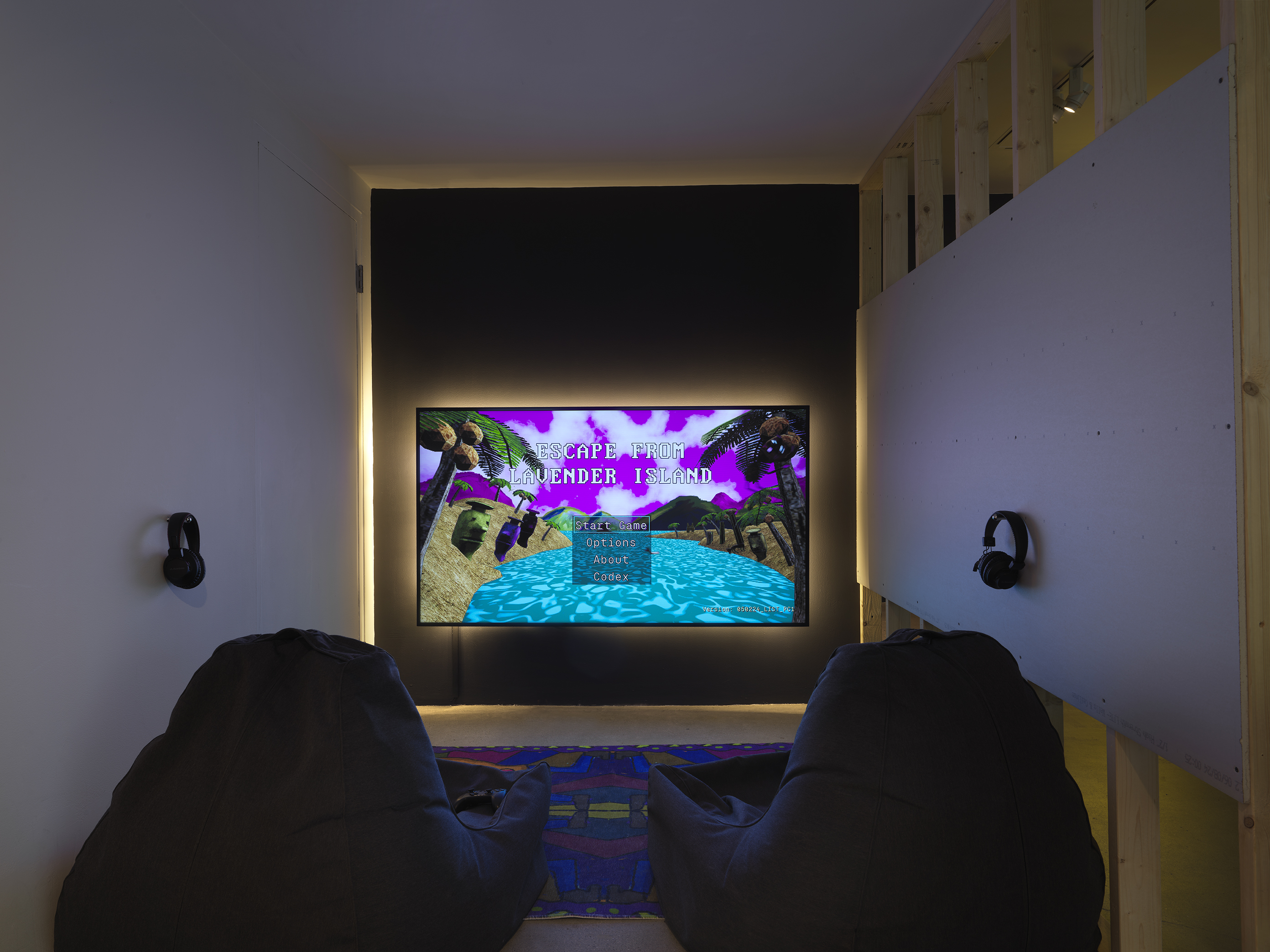 A backlit, glowing TV screen hangs on the wall close to the floor. A start menu  depicting a digitally rendered island scene for the videogame Escape from Lavender  Island is displayed on the TV. A pair of headphones hang off both the left and right  walls. Two soft chairs are placed on the floor over a colorful rug in front of the screen.