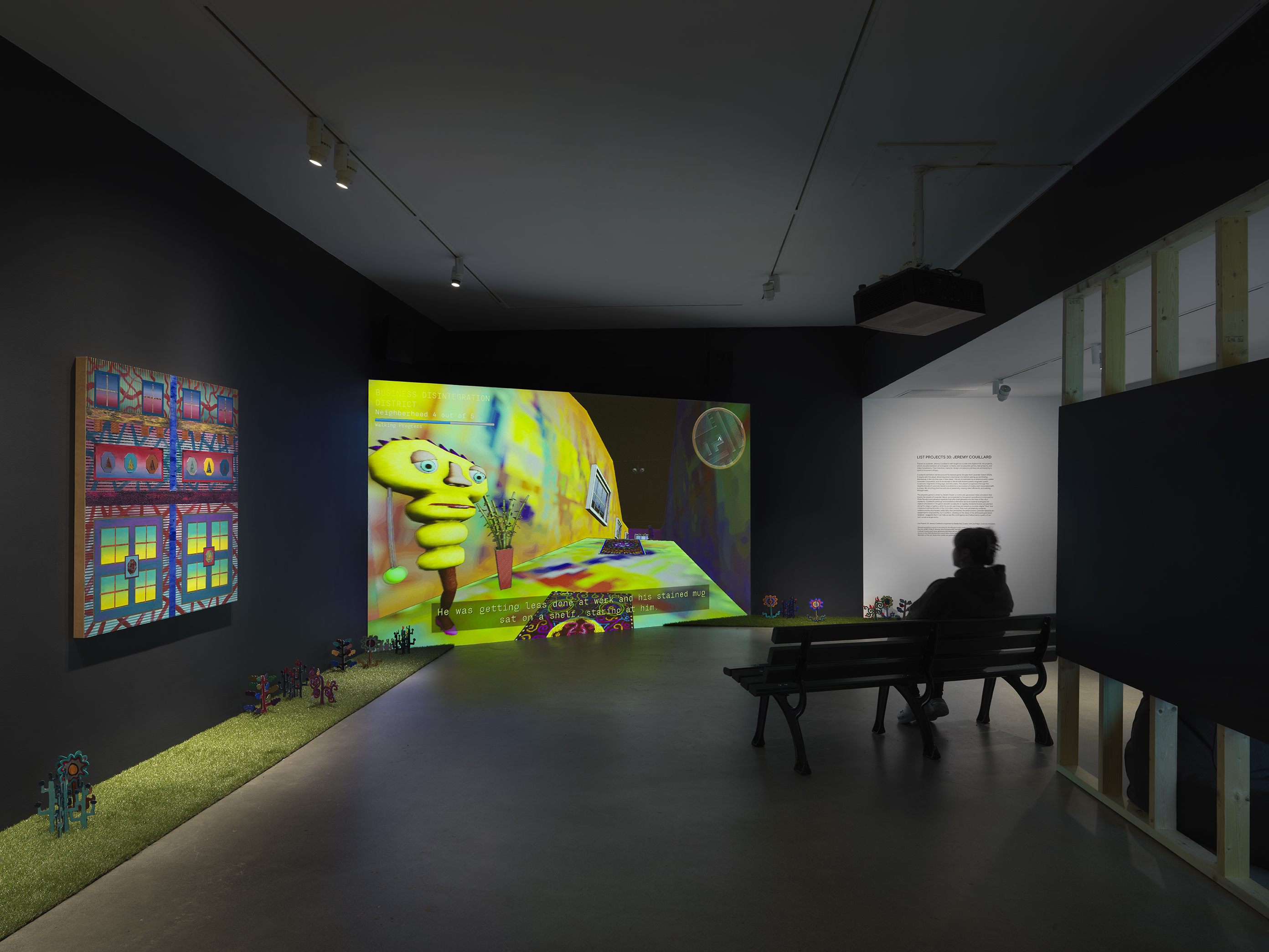 A dark, black-walled gallery space with artworks on display. On the left wall  hangs a painting depicting bright, geometric forms. In the back of the gallery, a large  screen illuminates the space displaying digitally rendered footage of a bright colored,  cartoon-like room. A woman sits on a park bench facing the screen. A strip of astroturf  lines the floor around the gallery, with painted wooden flowers emerging from it.