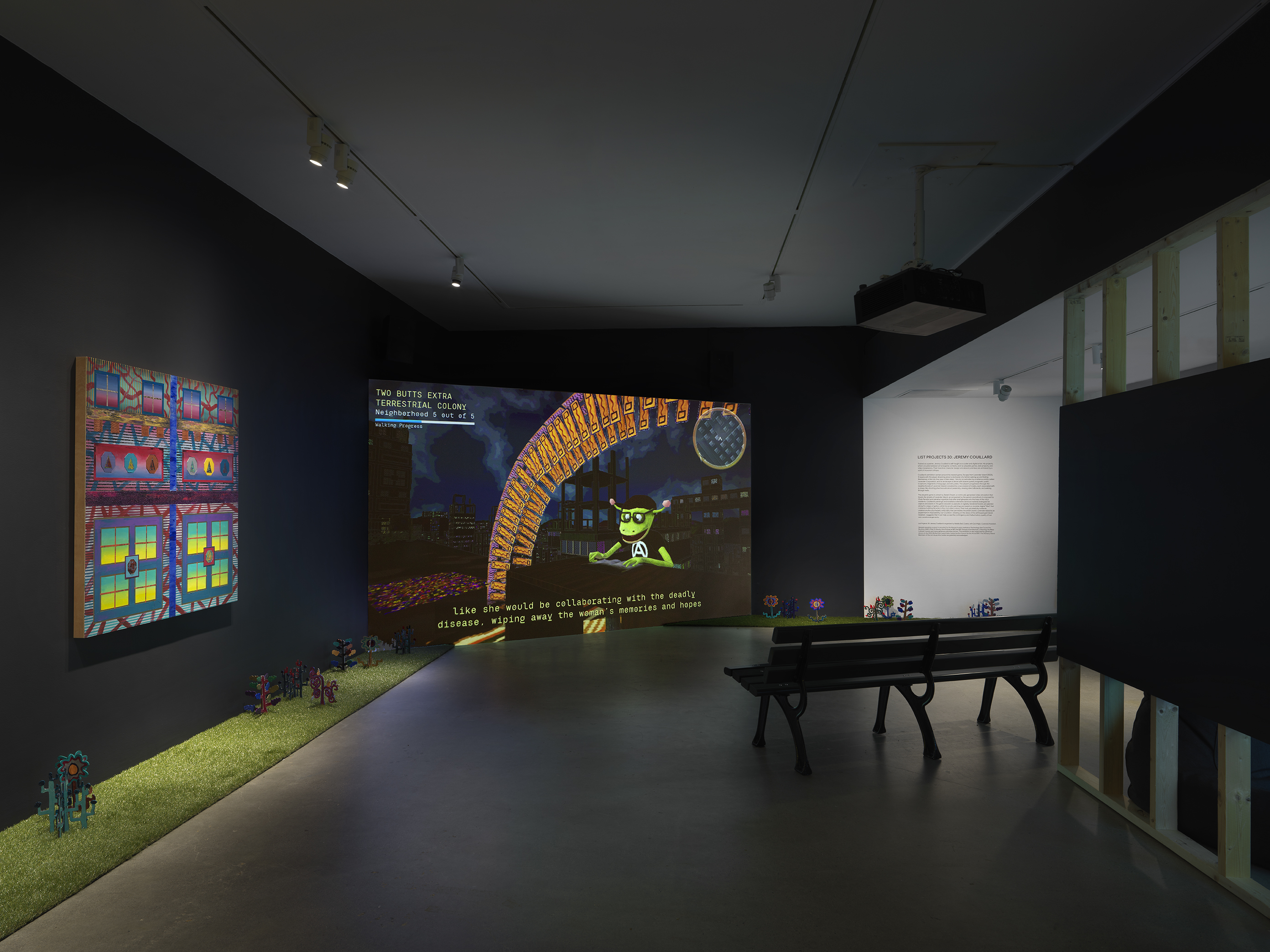 A dark, black-walled gallery space with artworks on display. On the left wall  hangs a painting depicting bright, geometric forms. In the back of the gallery, a large  screen illuminates the space displaying digitally rendered footage of a green creature  reading a book under an orange archway overlooking a city. A park bench faces the  screen. A strip of astroturf lines the floor around the gallery, with painted wooden flowers emerging from it. 