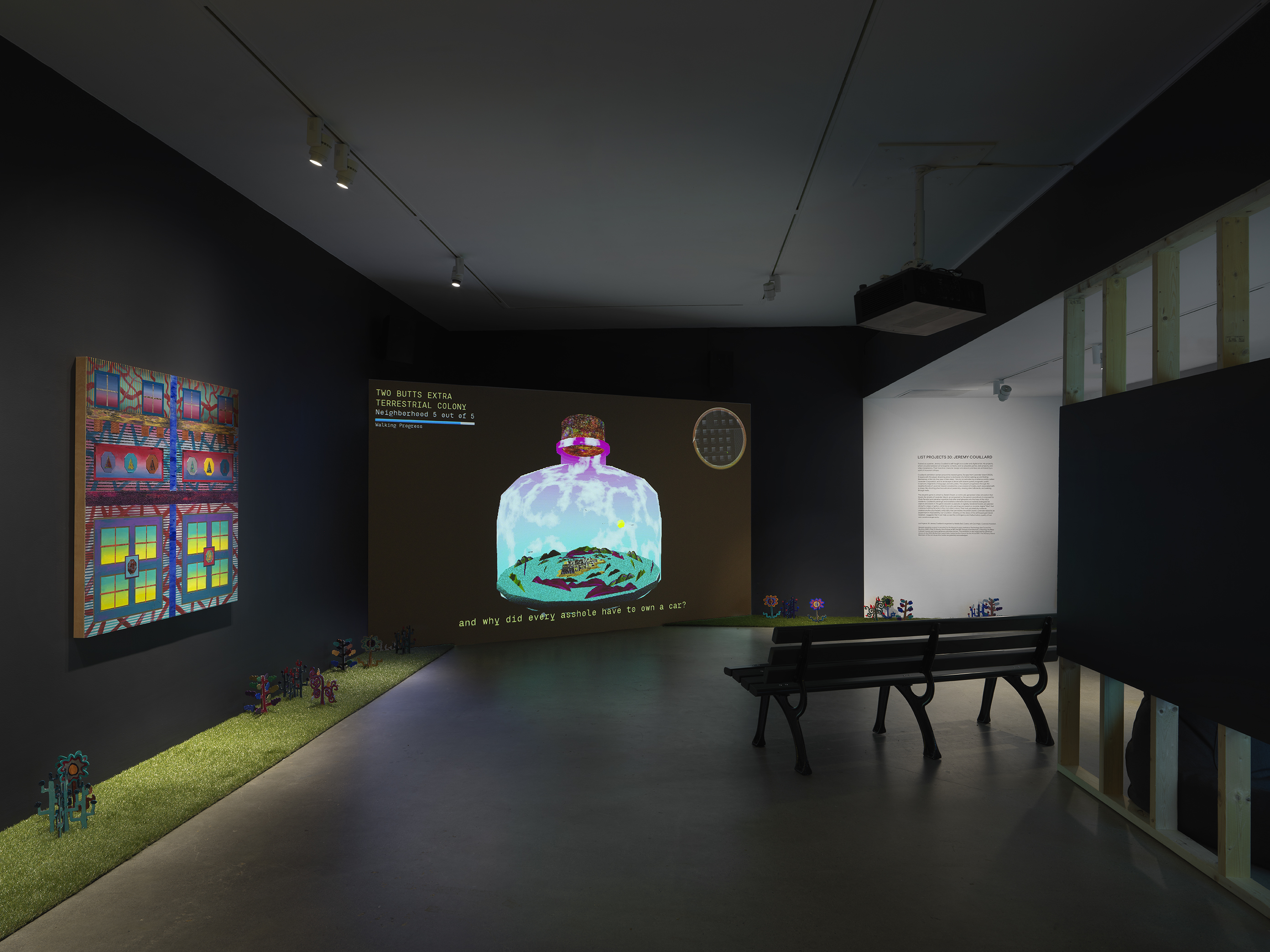 A dark, black-walled gallery space with artworks on display. On the left wall  hangs a painting depicting bright, geometric forms. In the back of the gallery, a large  screen illuminates the space displaying digitally rendered footage of a large bottle with  an island inside of it. A park bench faces the screen. A strip of astroturf lines the floor  around the gallery, with painted wooden flowers emerging from it.