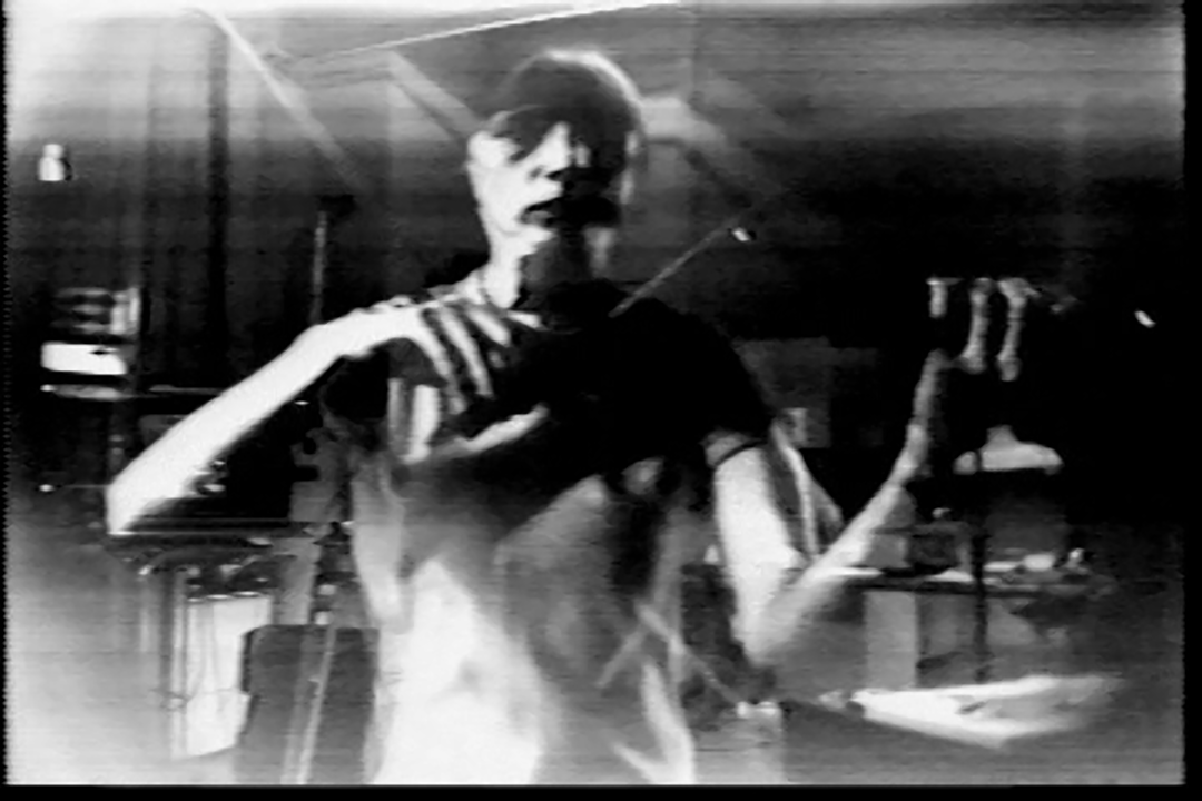 A woman plays a violin. She faces the viewer straight on. The image is grayscale and distinctly low resolution, overlayed with the ghost of another image