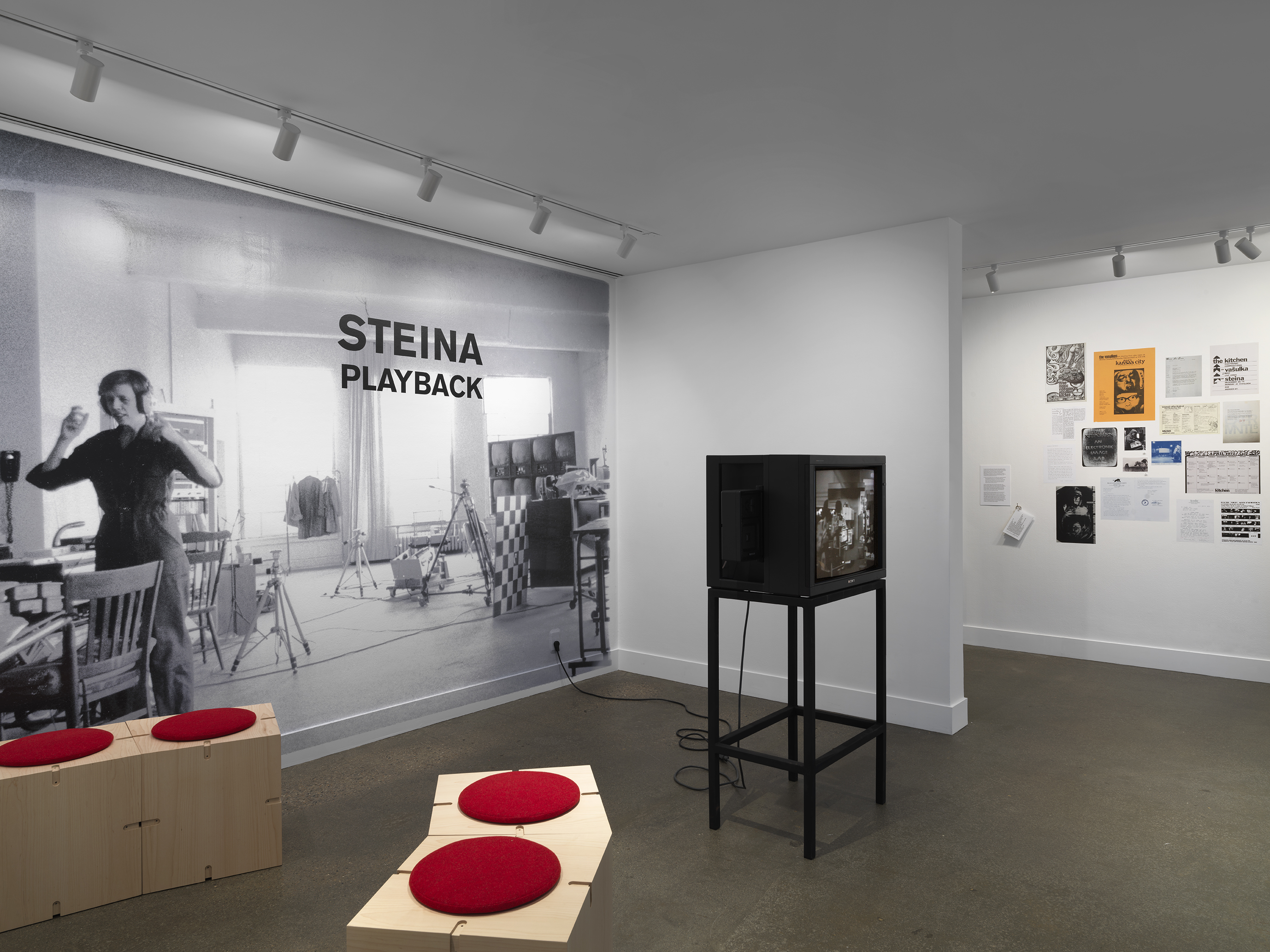 A black-and-white photograph printed mural-size on a wall shows a woman  wearing headphones. She is located in a studio full of technical equipment: tripods, video monitors, wiring. The photograph is overlaid with text indicating the exhibition title:  “Steina: Playback.” The gallery space also includes a boxy video monitor and wooden  seating. In the background on the right, posters and other documents are pasted on the wall. 