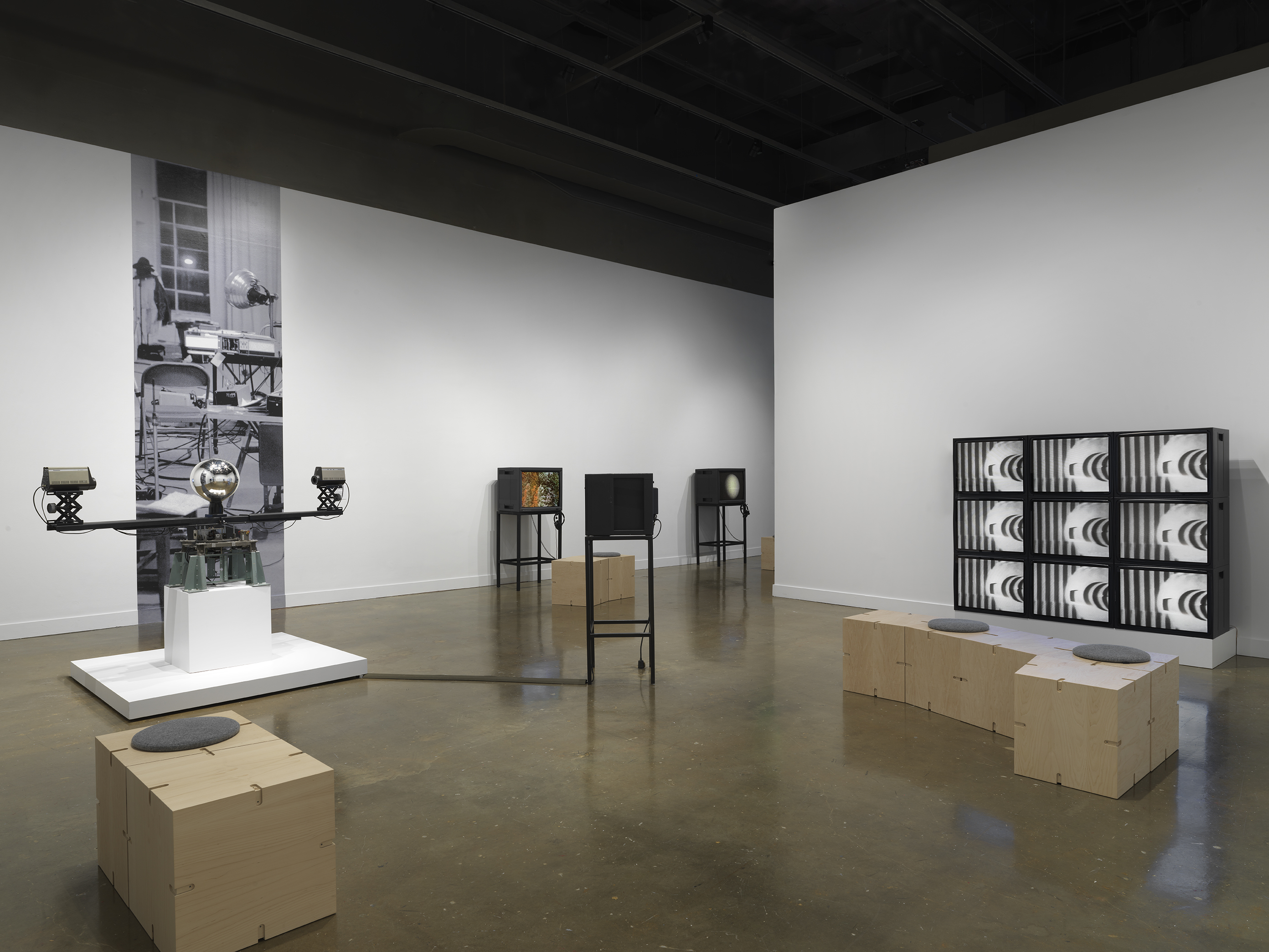 A gallery with several video works. At left is a sculptural apparatus with two  cameras on the ends of a single rotating arm, each pointed inwards at a central  mirrored sphere. At right is a grid of nine monitors, all showing the same abstract  black-and-white image. In the middle, three boxy video monitors on metal stands are visible in the background. 