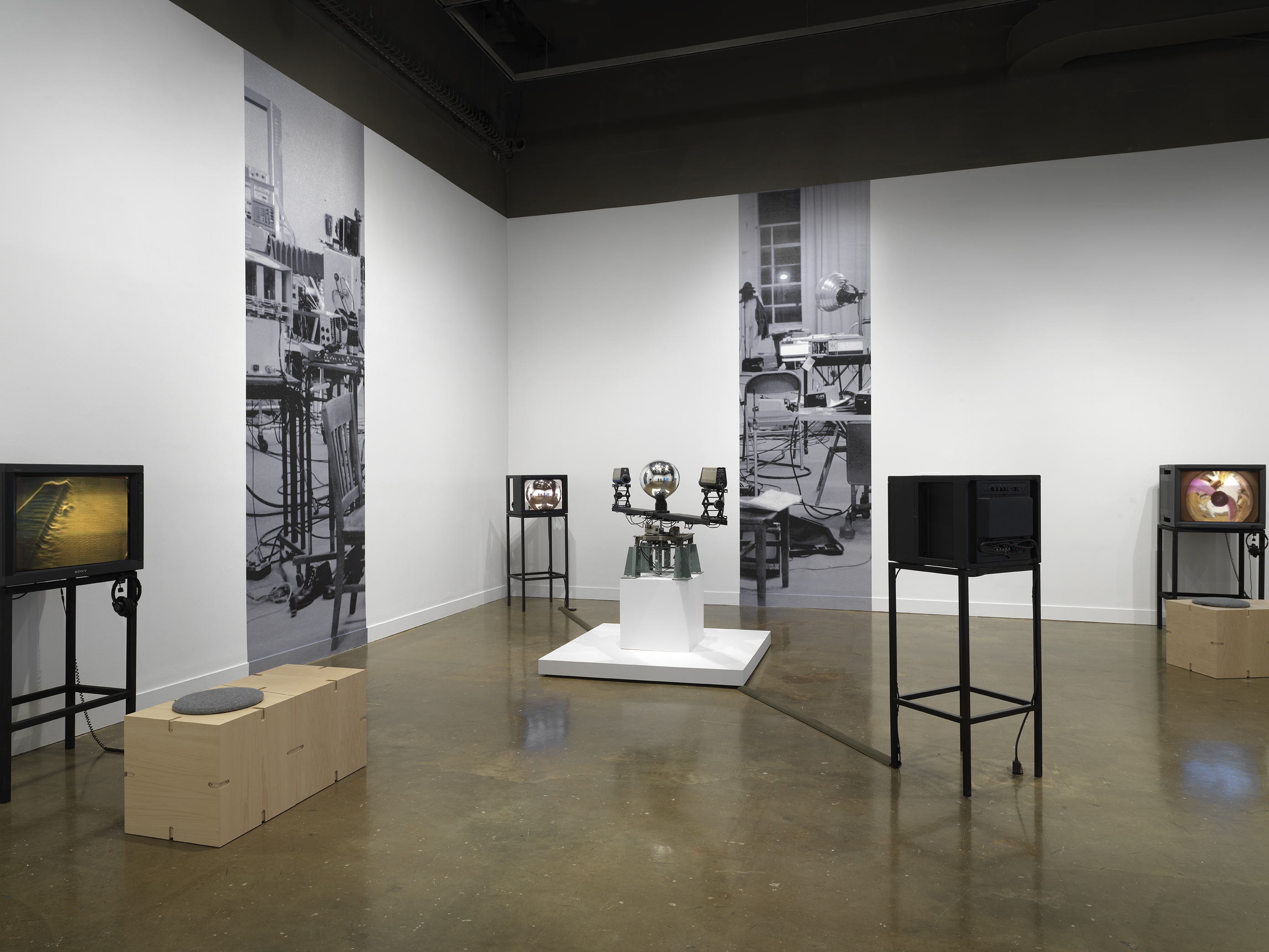 In the center, a sculptural apparatus comprises two cameras on the ends of a  single rotating arm, each pointed inwards at a central mirrored sphere. Feeds from the  cameras are routed to two monitors in the space. On the walls nearly, strips of a  photograph of a studio are pasted on the wall at large scale. On either edge of the  image, video monitors display other works; the one on the right shows a body distorted around a circle, recalling the feed of the orb. 