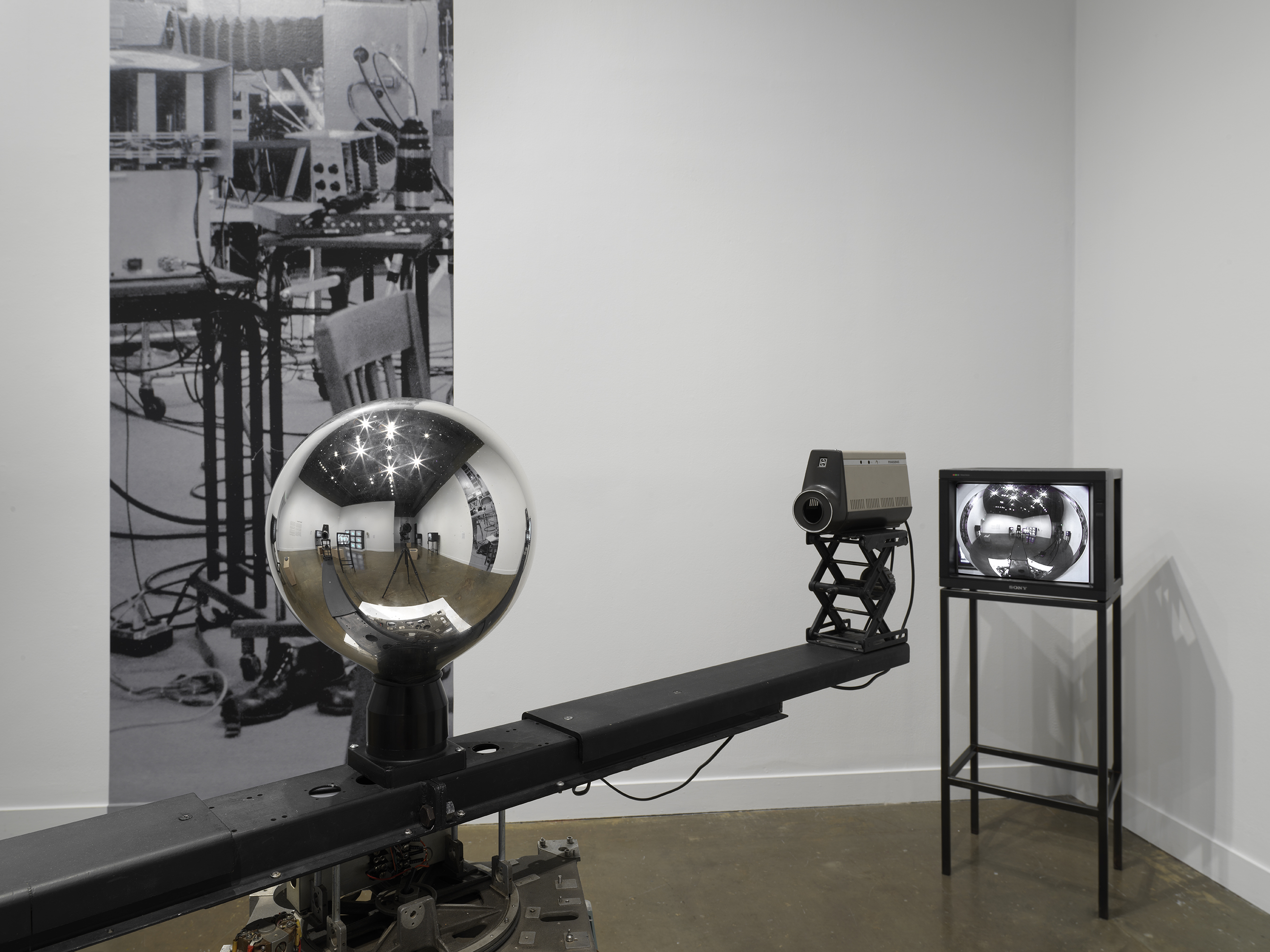 A mirrored sphere sits on top of a rotating central arm with cameras at each  end. In this close-up view, we see the details of the room within the orb: video monitors,  lights, the tripod taking the photo. In the corner behind the sculpture, a monitor shows a  feed of the orb, within which details of the room are also faintly visible. On the wall is a  narrow strip of a photograph of a studio space filled with technical equipment.
