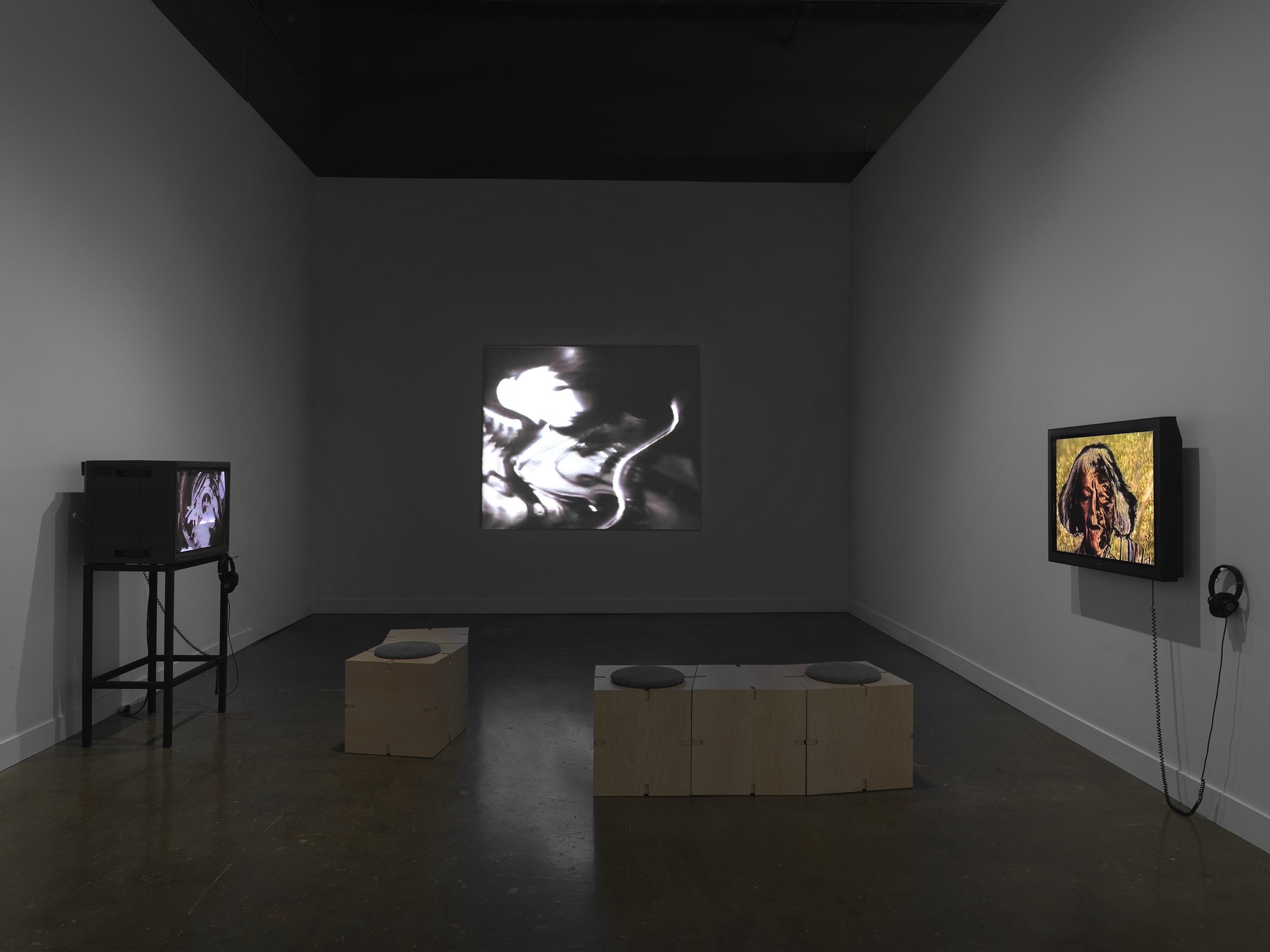 Three videos are shown in a gallery space. At center, a distorted image of a‬ ‭ person playing the violin is projected on the wall: the bow is curved and the person‬ ‭ indistinct, hinting at its electronic manipulation. At right, a flat monitor shows an image of‬ ‭ an older woman against a yellow background; portions of the background show through‬ ‭the face. At right, a boxy video monitor sits on a stand.‬ ‭