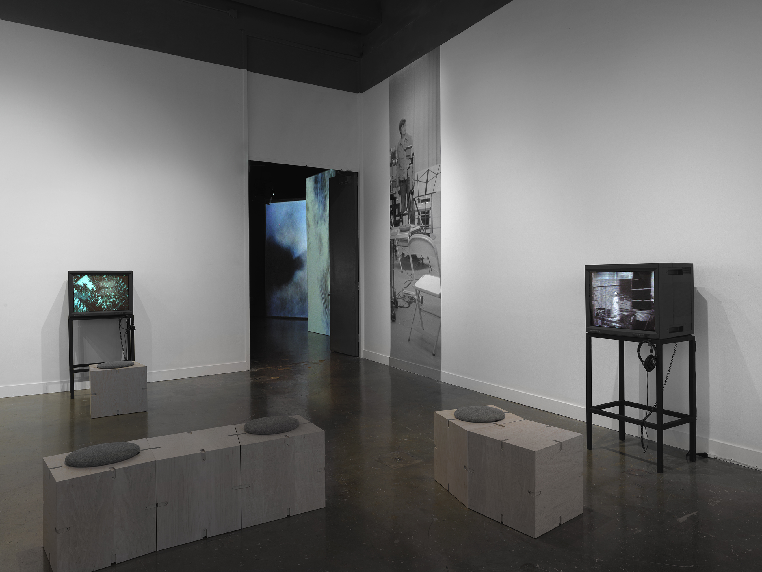 A gallery contains two boxy video monitors on metal stands, each with an  accompanying pair of headphones. The monitor on the left shows a color image that  could be the outline of trees against the sky. The right monitor shows a black-and-white  image of boxy shapes, possibly electronic equipment. A narrow slice of an image is  printed on the wall in the background, showing a woman in a studio space. Further  back, through a doorway, we glimpse a darker gallery that contains large-scale video 