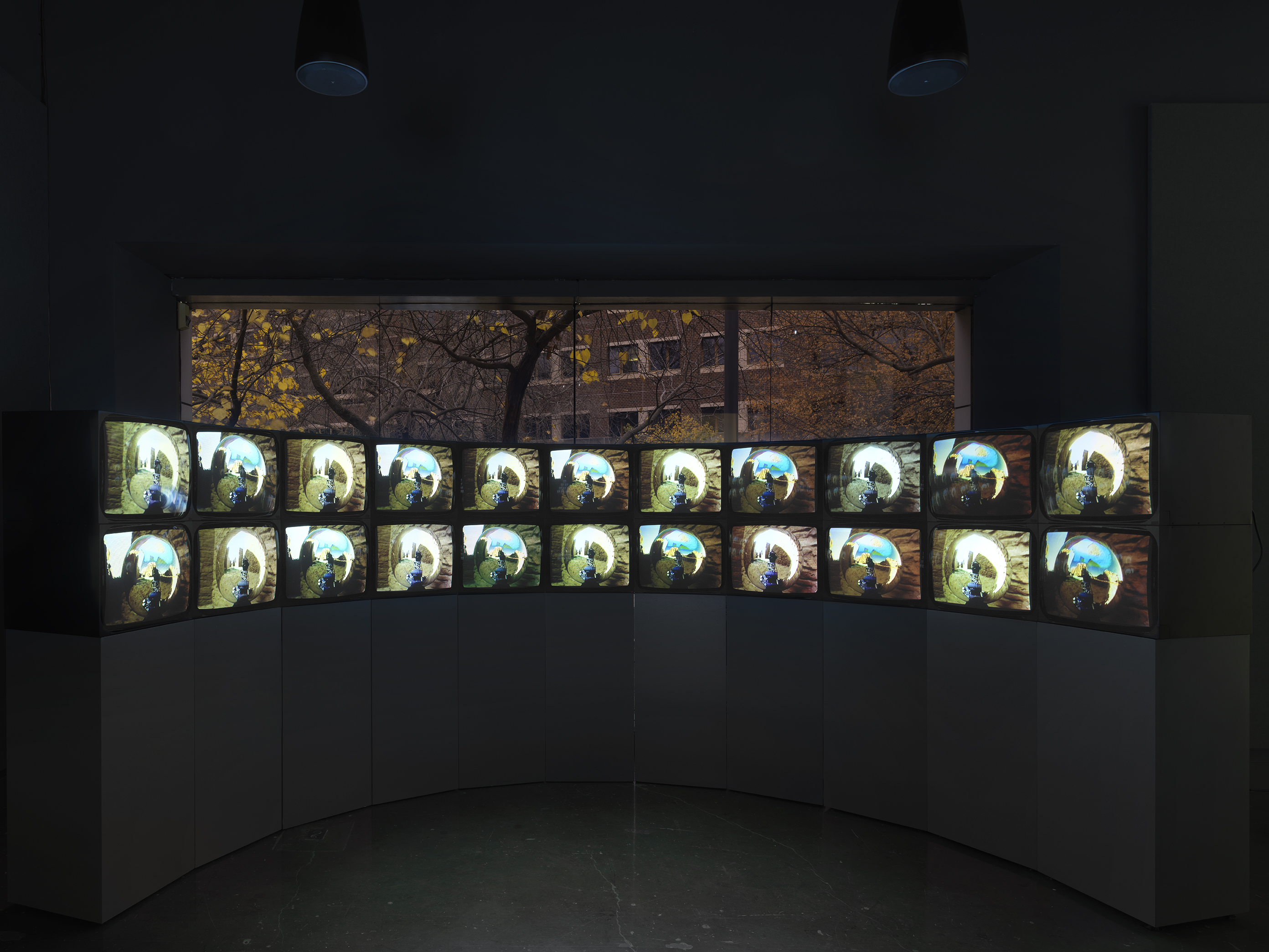 A double-stacked arc of boxy video monitors sits on a pedestal in a darkened  gallery; there are 22 in all. The monitors show similar but not identical images: in each,  a central mirrored orb reflects and distorts the surrounding environment, which includes  a stone wall and blue sky. Behind the video installation is a large window, through which  trees and another building can be seen.