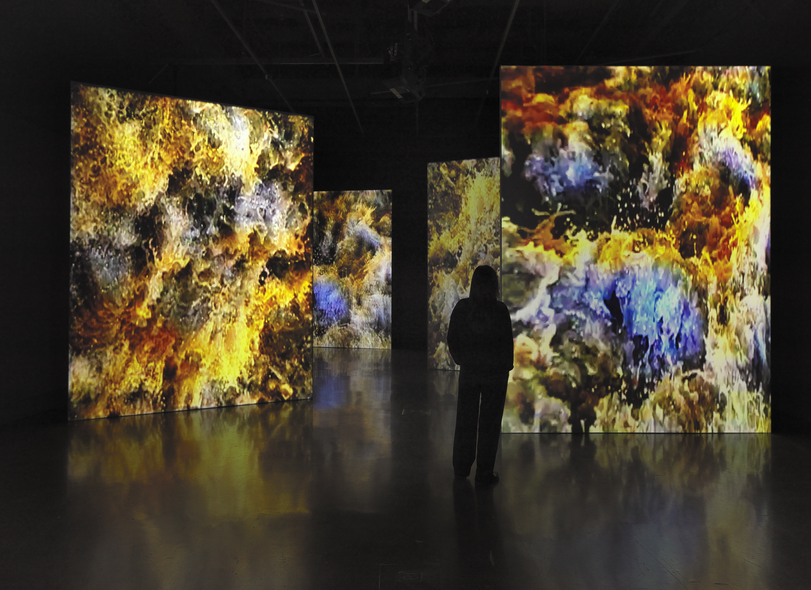 Four large vertically oriented video screens fill a dark gallery space. Each of  them shows a close-up image of water, captured in a state of frozen turbulence and in saturated shades of blue and yellow. In the foreground, a person in the gallery is silhouetted against the video projections. 