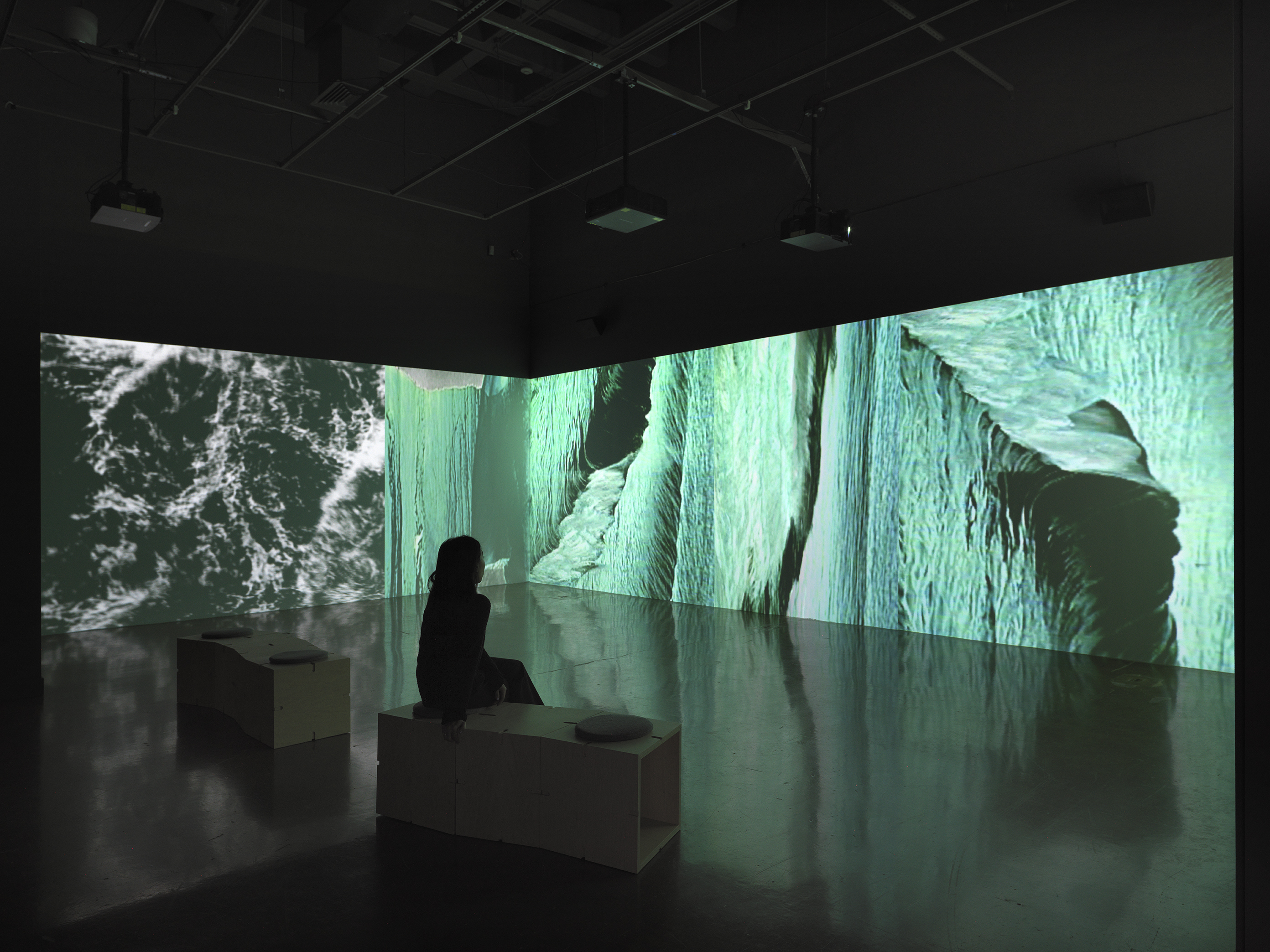 A continuous surface of projected video fills a wall and wraps around a corner  in a dark gallery. The images are liquid-like semi-abstractions of blue-green, white, and  black, recalling the surface of moving water. A person sits on one of two wooden  benches that are faintly visible in the foreground.