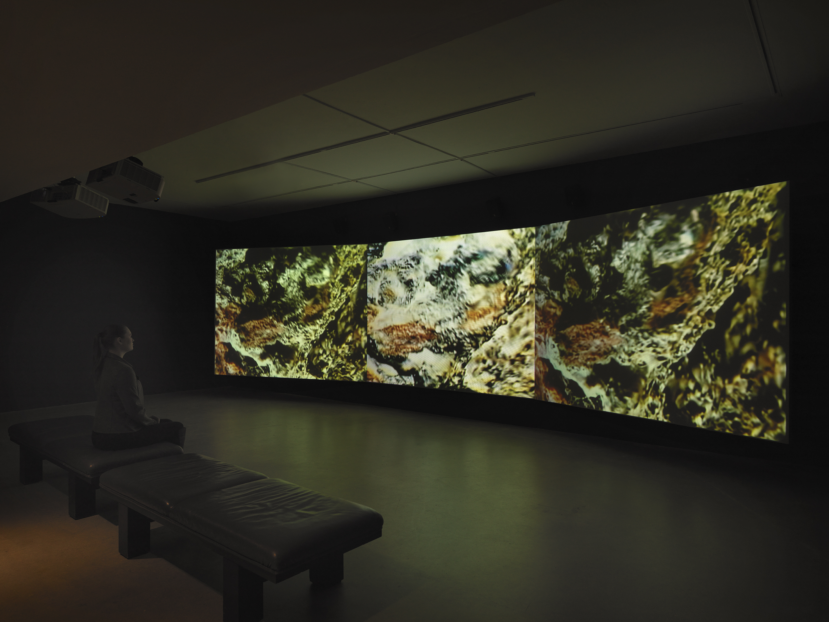 Three video projections fill the wall of a dark gallery space: each shows a textural yet topographical pattern that might be a close shot of earth or stone. Two  benches are faintly visible in the left foreground; a person sits on one and watches the video. 