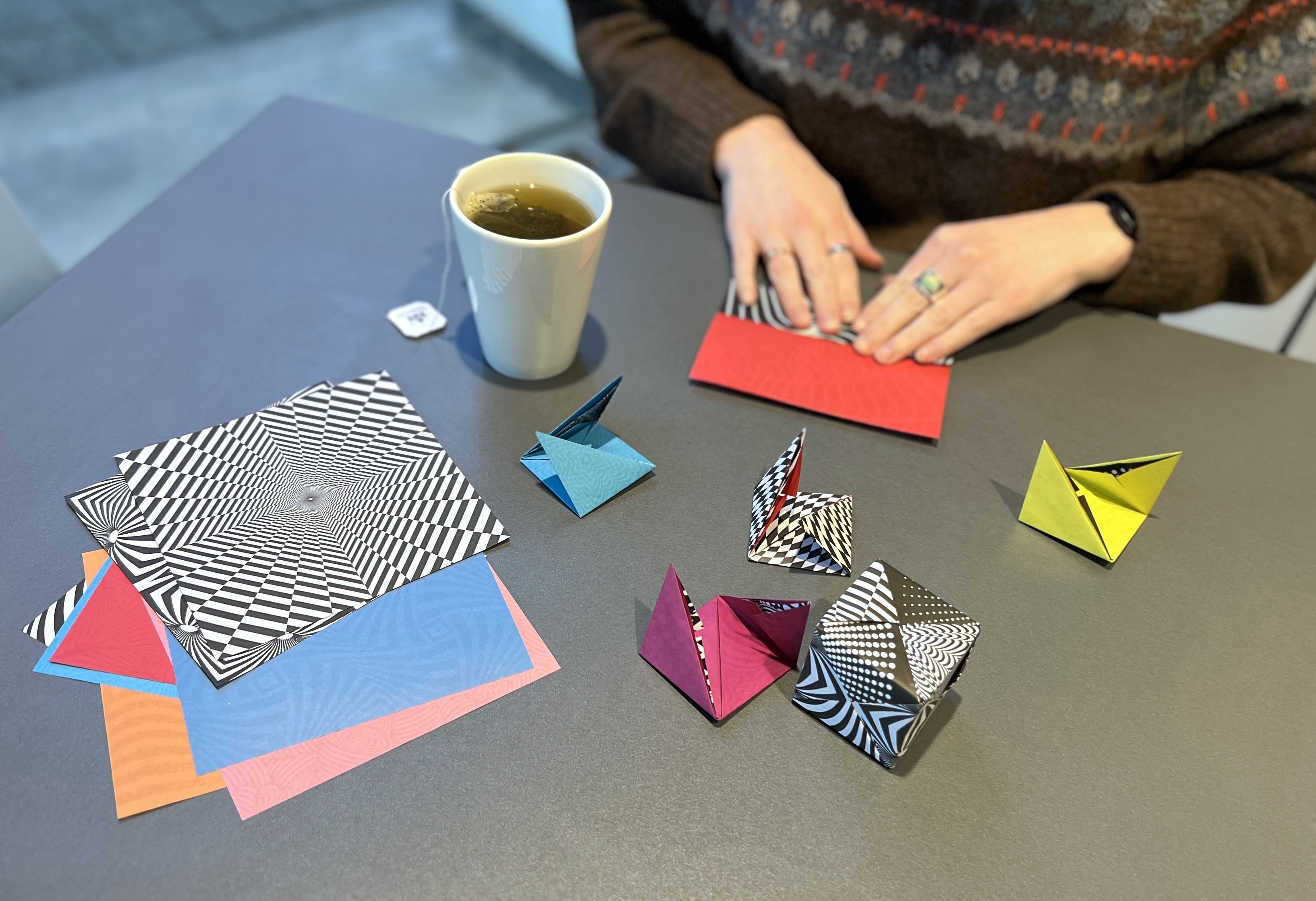 Two hands folding colorful geometric paper. There is a white mug of tea to the left side of the hands and a stack of unfolded paper in the foreground.