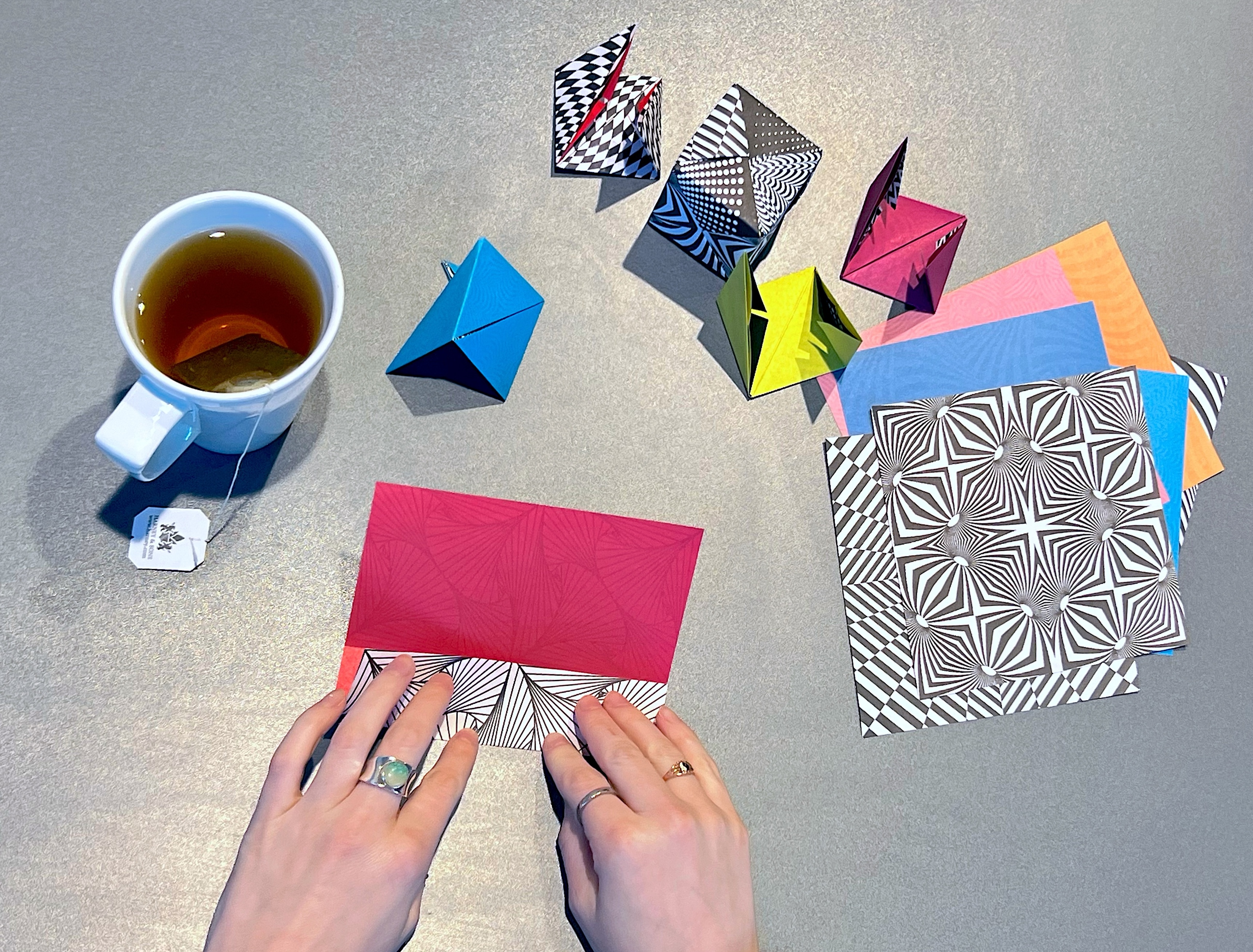 Overhead view of two hands folding colorful geometric paper. There is a white mug of tea to the left side and a stack of unfolded paper to the right.