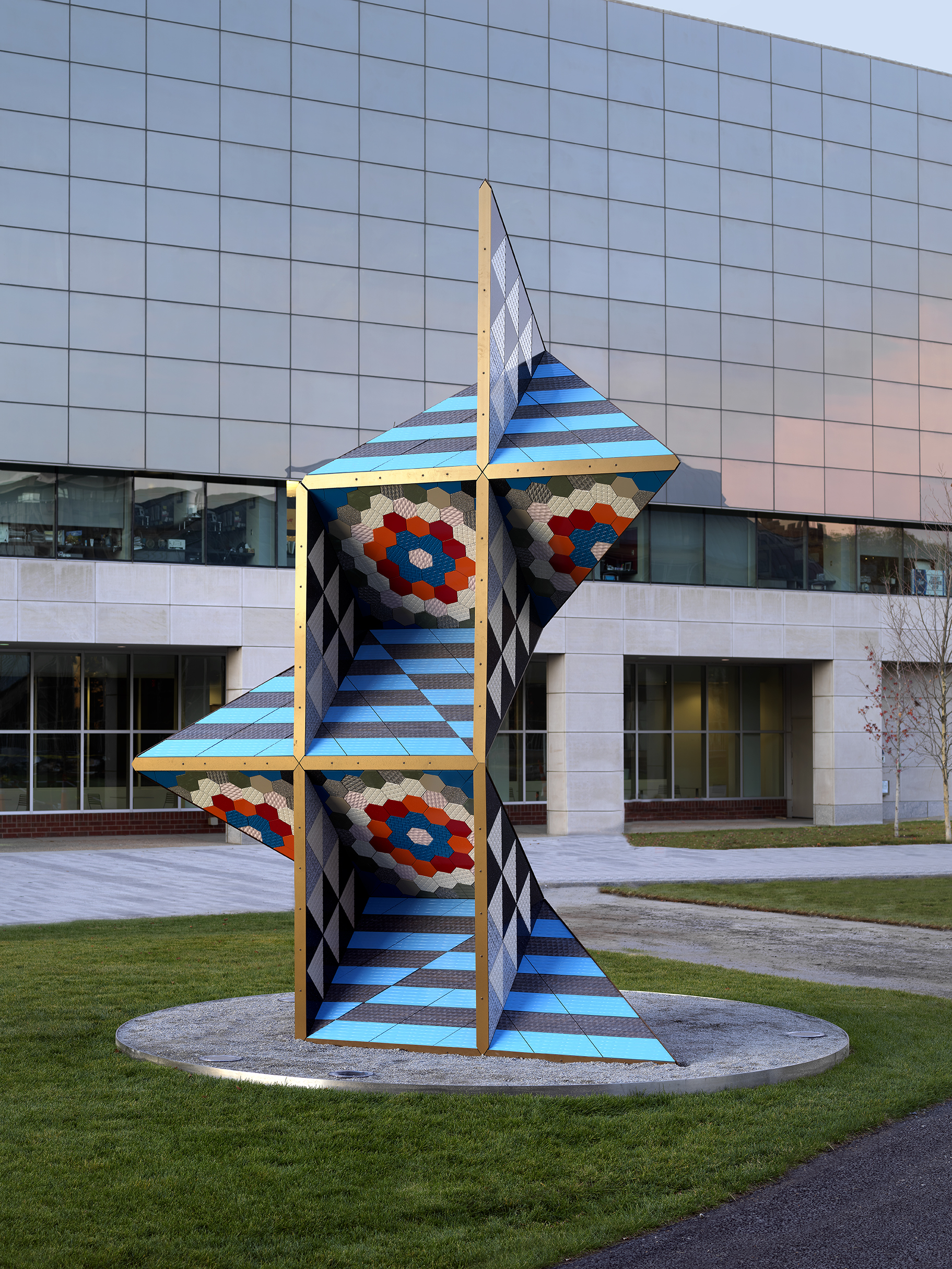 Large brightly colored geometric sculpture in the middle of a grass lawn in front of a concrete building with reflective blue glass windows.