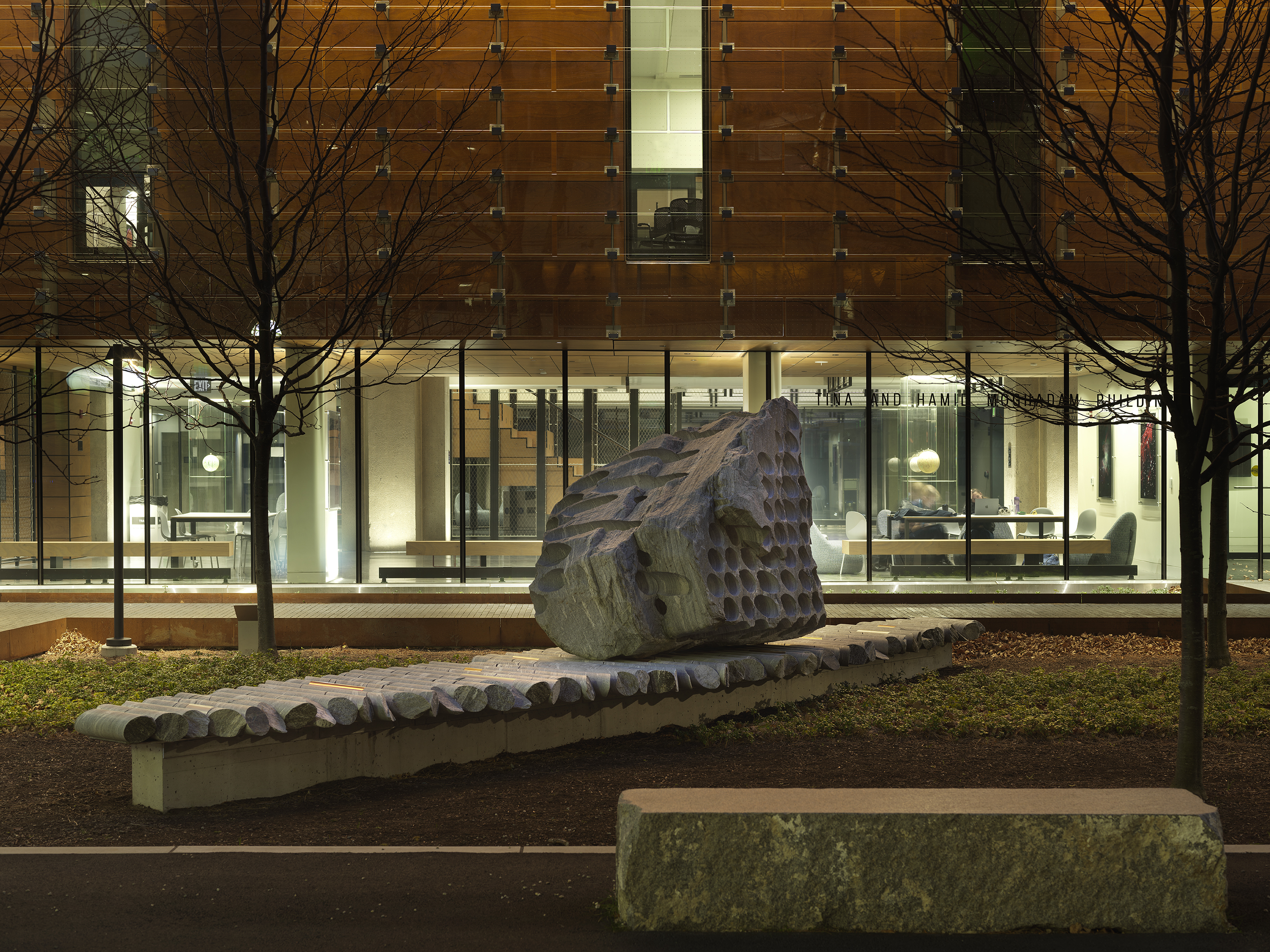 A large rock sculpture rests on a long strip of stone cylinders in a green space next to the corner of a building at night. The rock has a series of holes removed from its core, with the cylinders that were removed making up the strip it rests on.