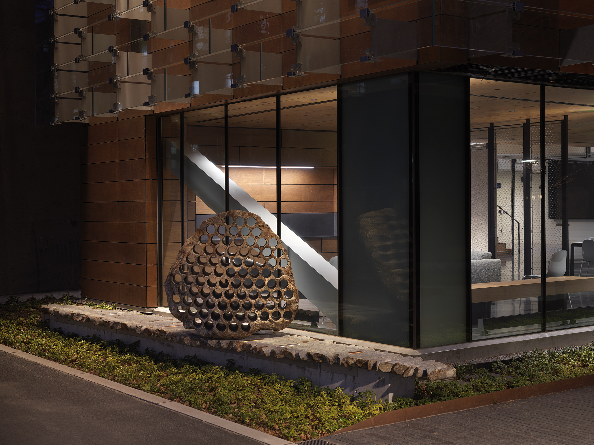 A large rock sculpture rests on a long strip of stone cylinders in a green space next to the corner of a building at night. The rock has a series of holes removed from its core, with the cylinders that were removed making up the strip it rests on.