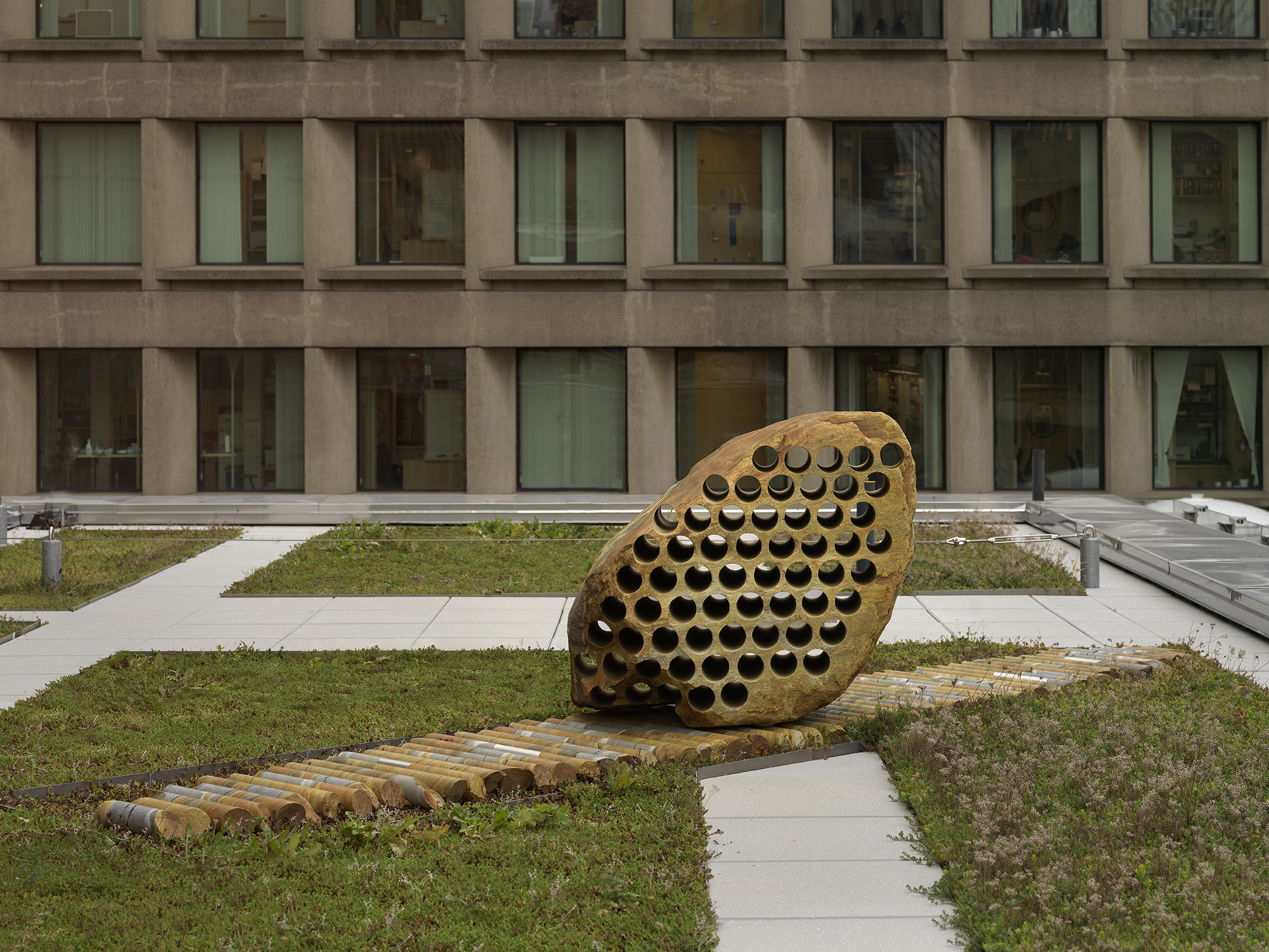 A large rock sculpture rests on a long strip of stone cylinders in a greenspace. The rock has a series of holes removed from its core, with the cylinders that were removed making up the strip it rests on. There is a pathway behind the sculpture and building tops surrounding the open space.