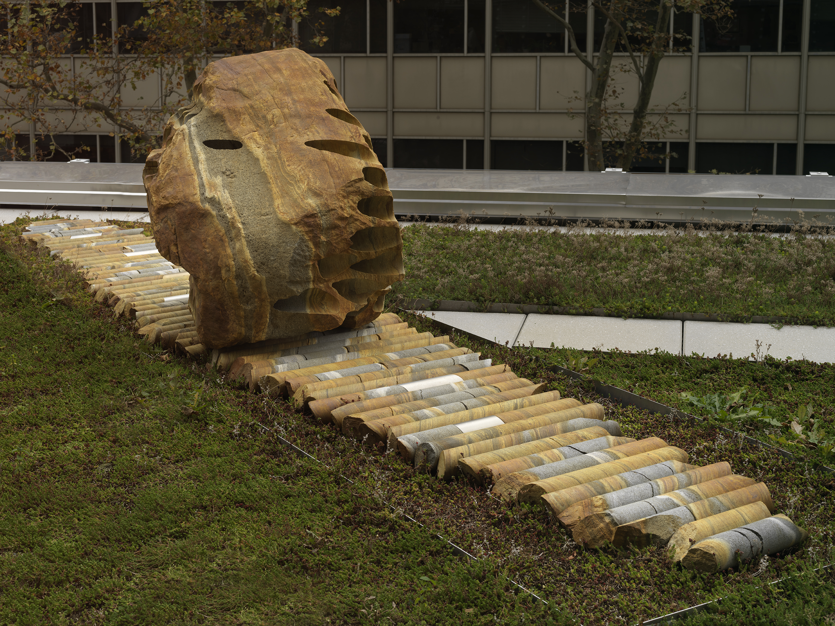A large rock sculpture rests on a long strip of stone cylinders in a greenspace. The rock has a series of holes removed from its core, with the cylinders that were removed making up the strip it rests on.