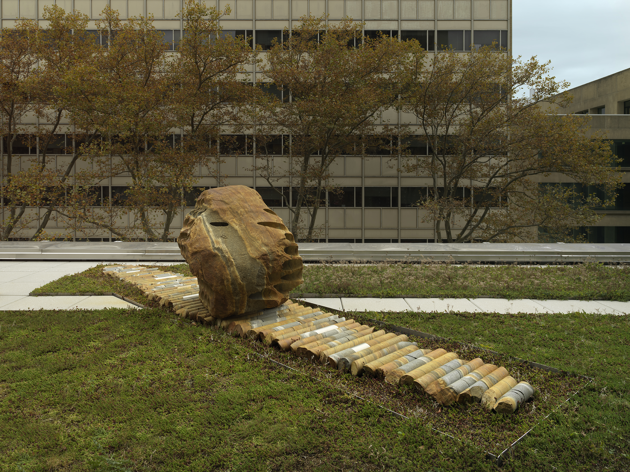 A large rock sculpture rests on a long strip of stone cylinders in a greenspace. The rock has a series of holes removed from its core, with the cylinders that were removed making up the strip it rests on. There is a pathway behind the sculpture and building tops surrounding the open space.