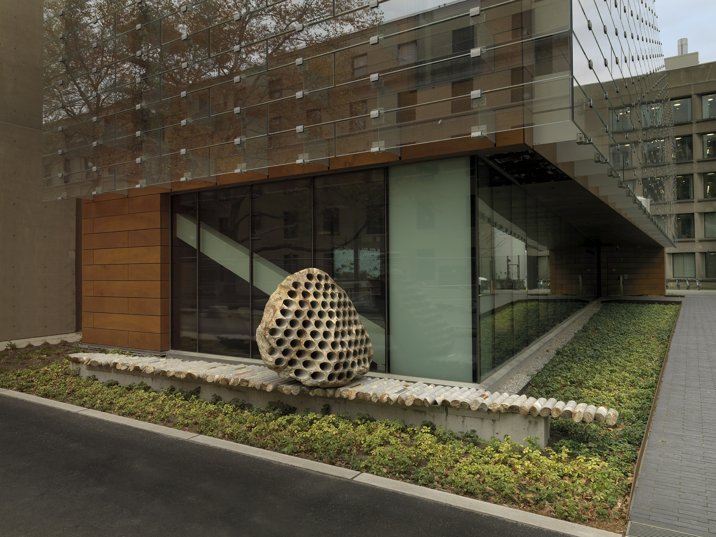 A large rock sculpture rests on a long strip of stone cylinders in a  greenspace next to the corner of a building. The rock has a series of holes  removed from its core, with the cylinders that were removed making up the strip it rests on.