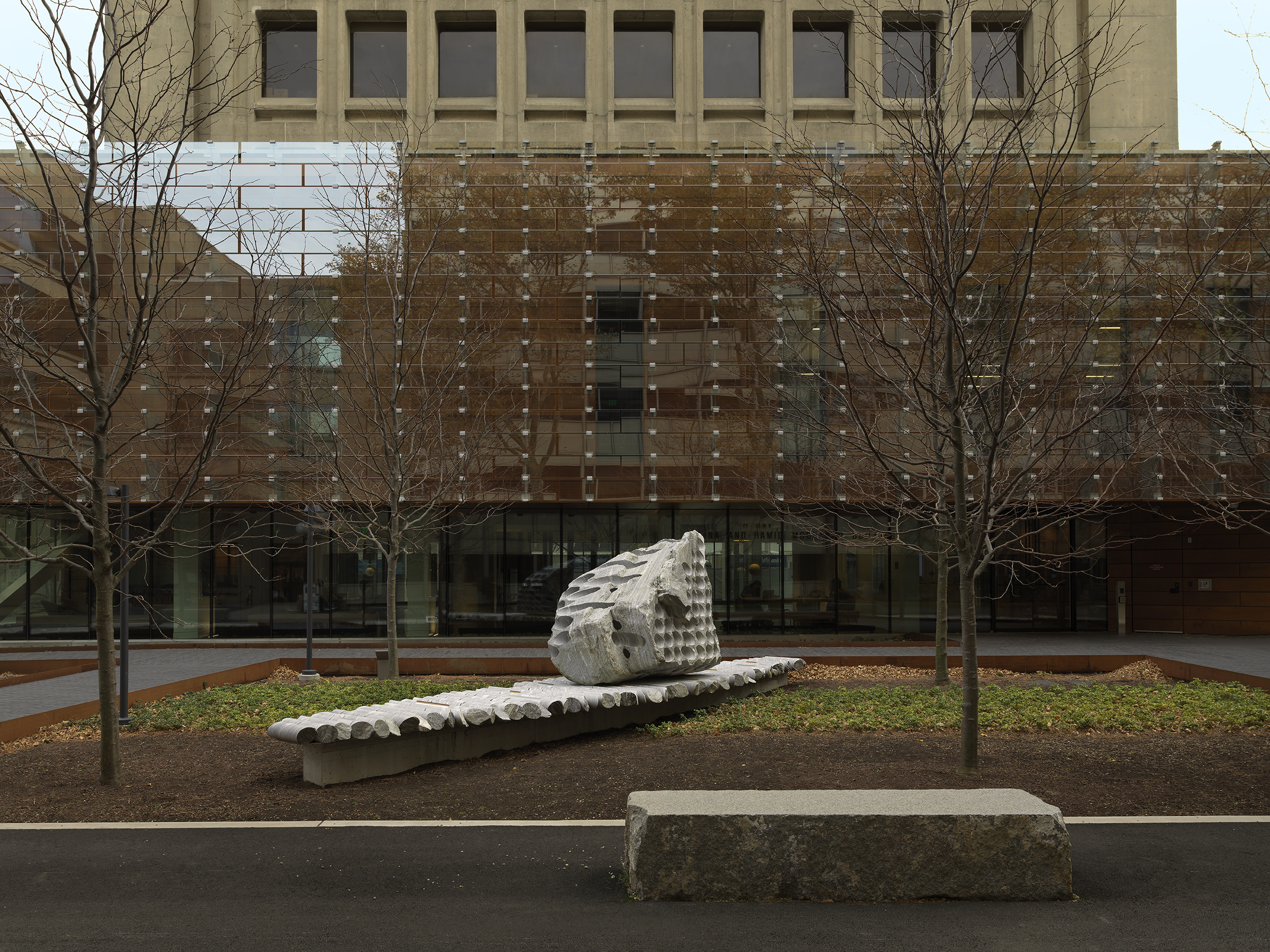 A large rock sculpture rests on a long strip of stone cylinders in an open space. The rock has a series of holes removed from its core, with the cylinders that were removed making up the strip it rests on. Trees, a pathway, and a gray building reside in the background. 