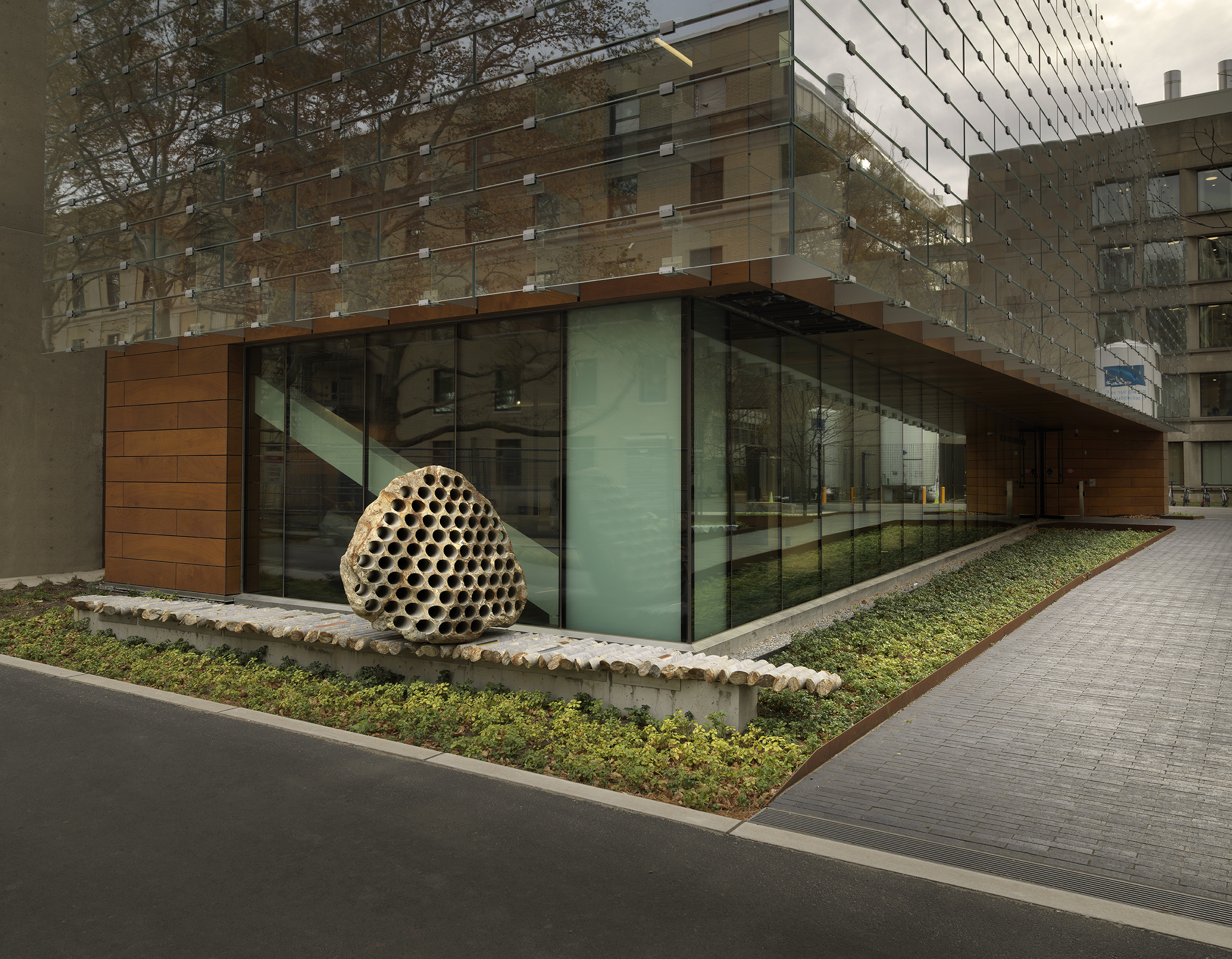 A large rock sculpture rests on a long strip of stone cylinders in a  greenspace next to the corner of a building. The rock has a series of holes  removed from its core, with the cylinders that were removed making up the strip it rests on. 