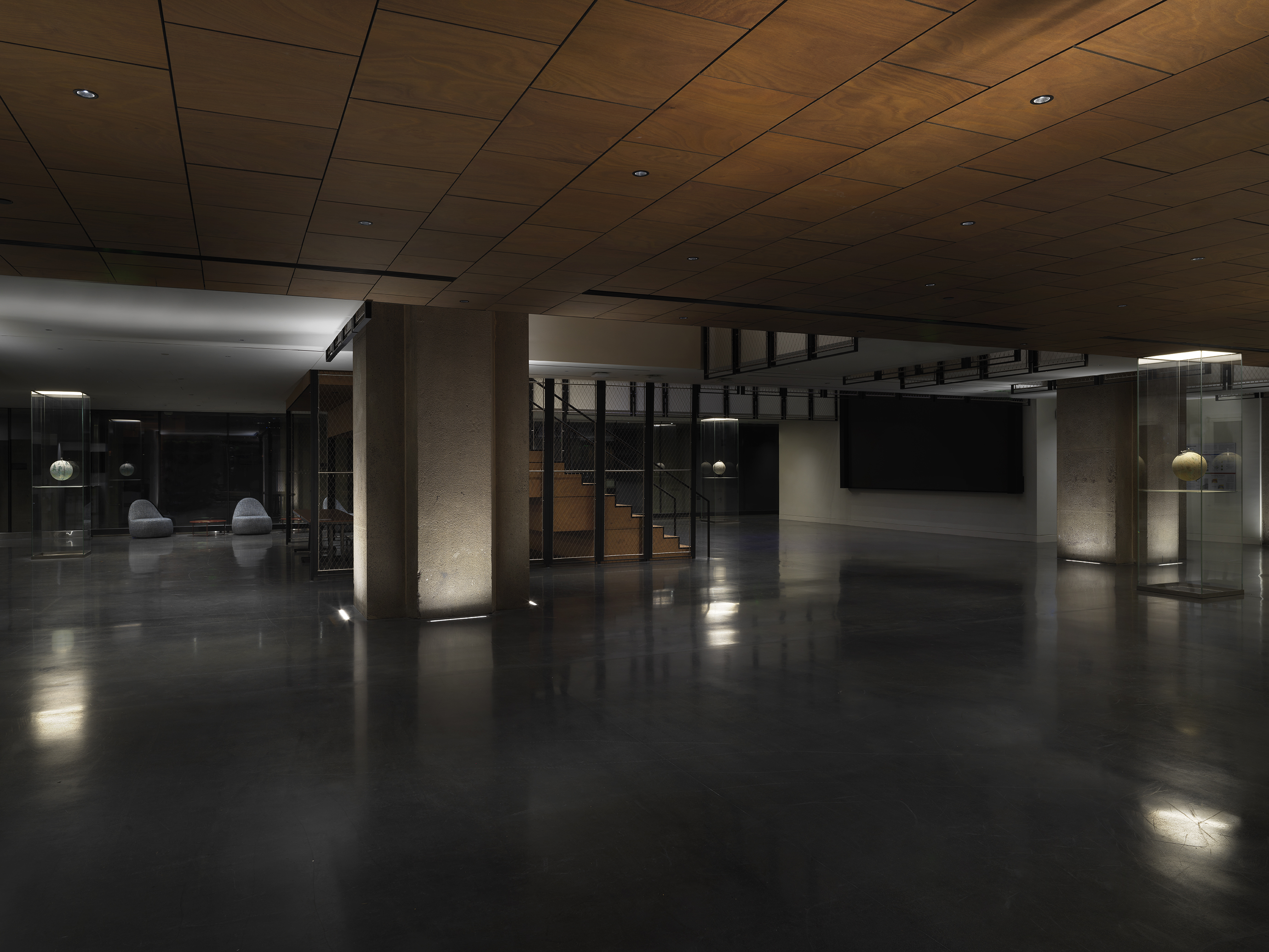 Three painted globe suspended in a glass rectangular columns can be seen throughout an open lobby. There are two gray chairs on the left hand side and a large wooden staircase in the center of the lobby faces a large digital screen hung on the right hand wall.
