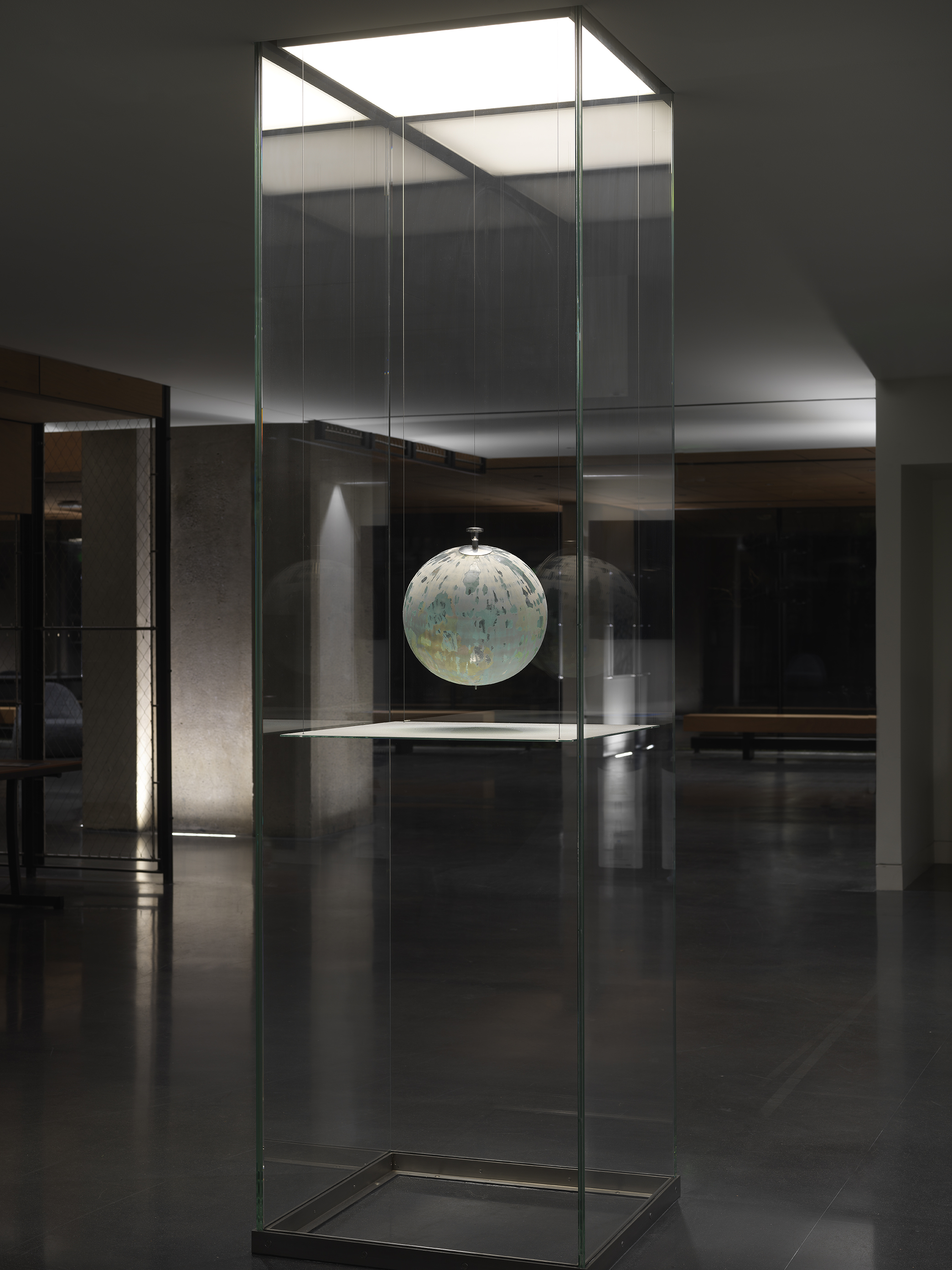 One painted globe suspended in a glass rectangular column and hangs over a flat surface that cuts the cylinder horizontally. Splatters of paint can be seen on top of the panel underneath the globe.