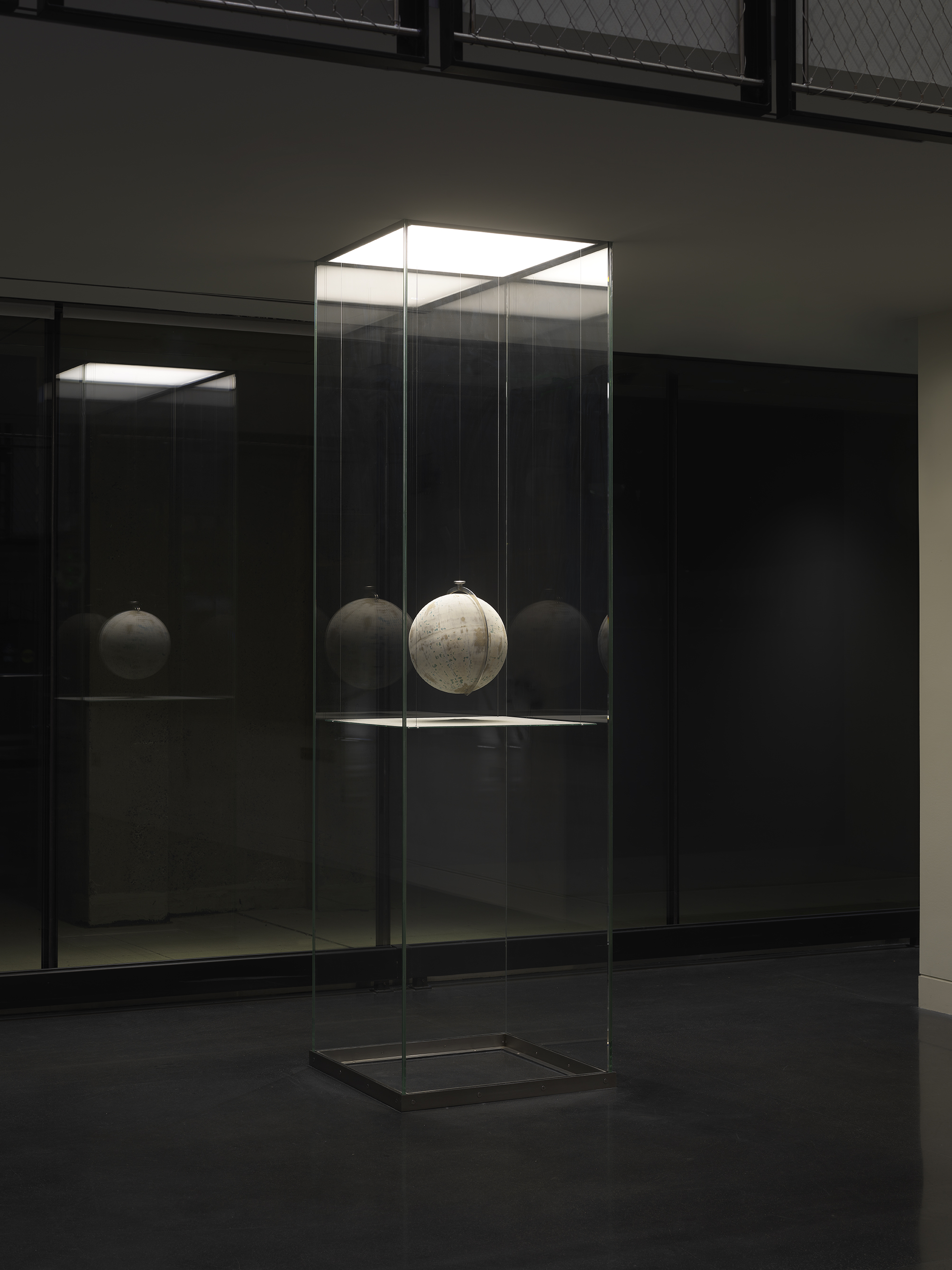 One painted globe suspended in a glass rectangular column and hangs over a flat surface that cuts the cylinder horizontally. Splatters of paint can be seen on top of the panel underneath the globe.