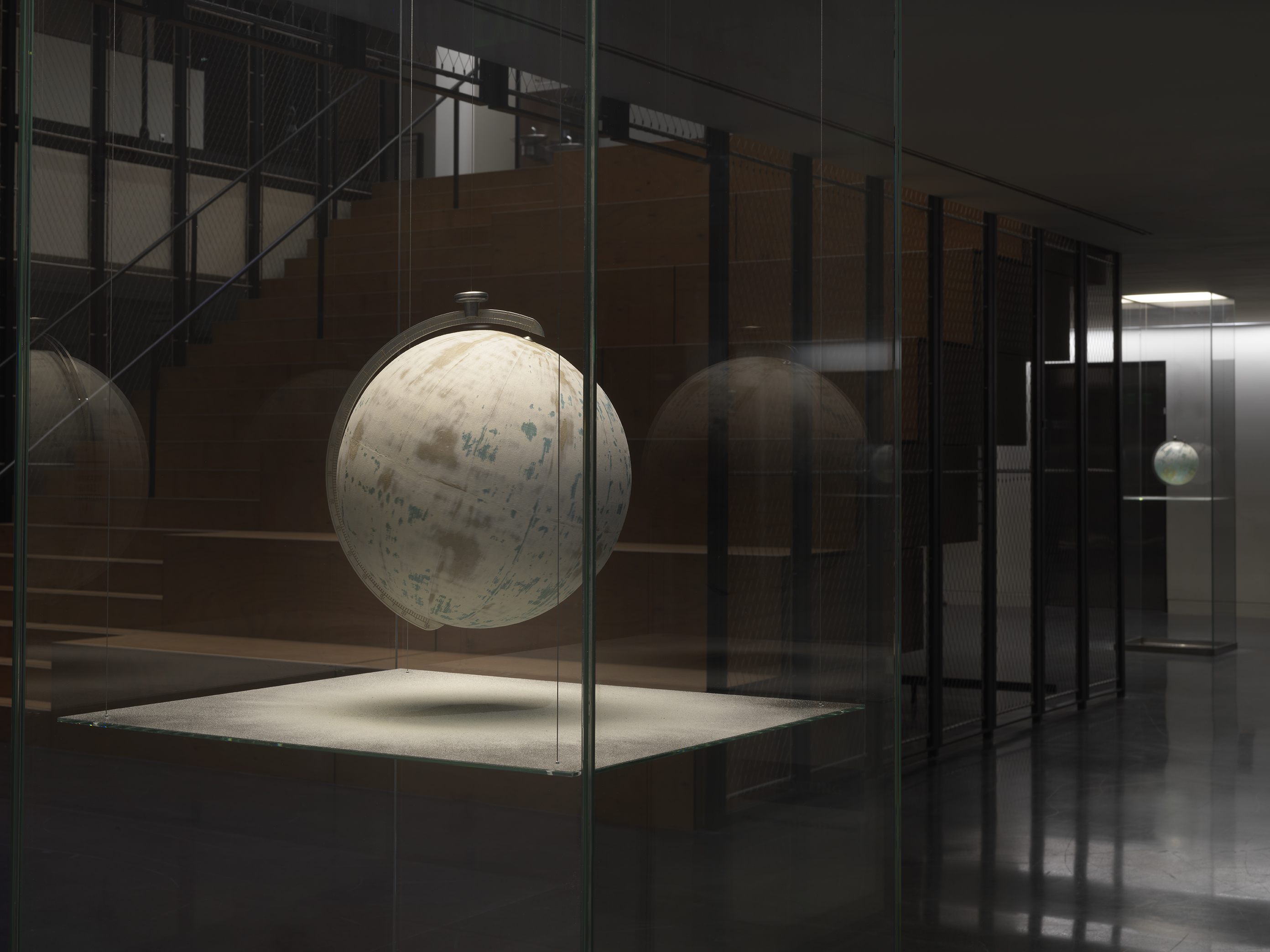 One painted globe suspended in a glass rectangular column and hangs over a flat surface that cuts the cylinder horizontally. Splatters of paint can be seen on top of the panel underneath the globe.