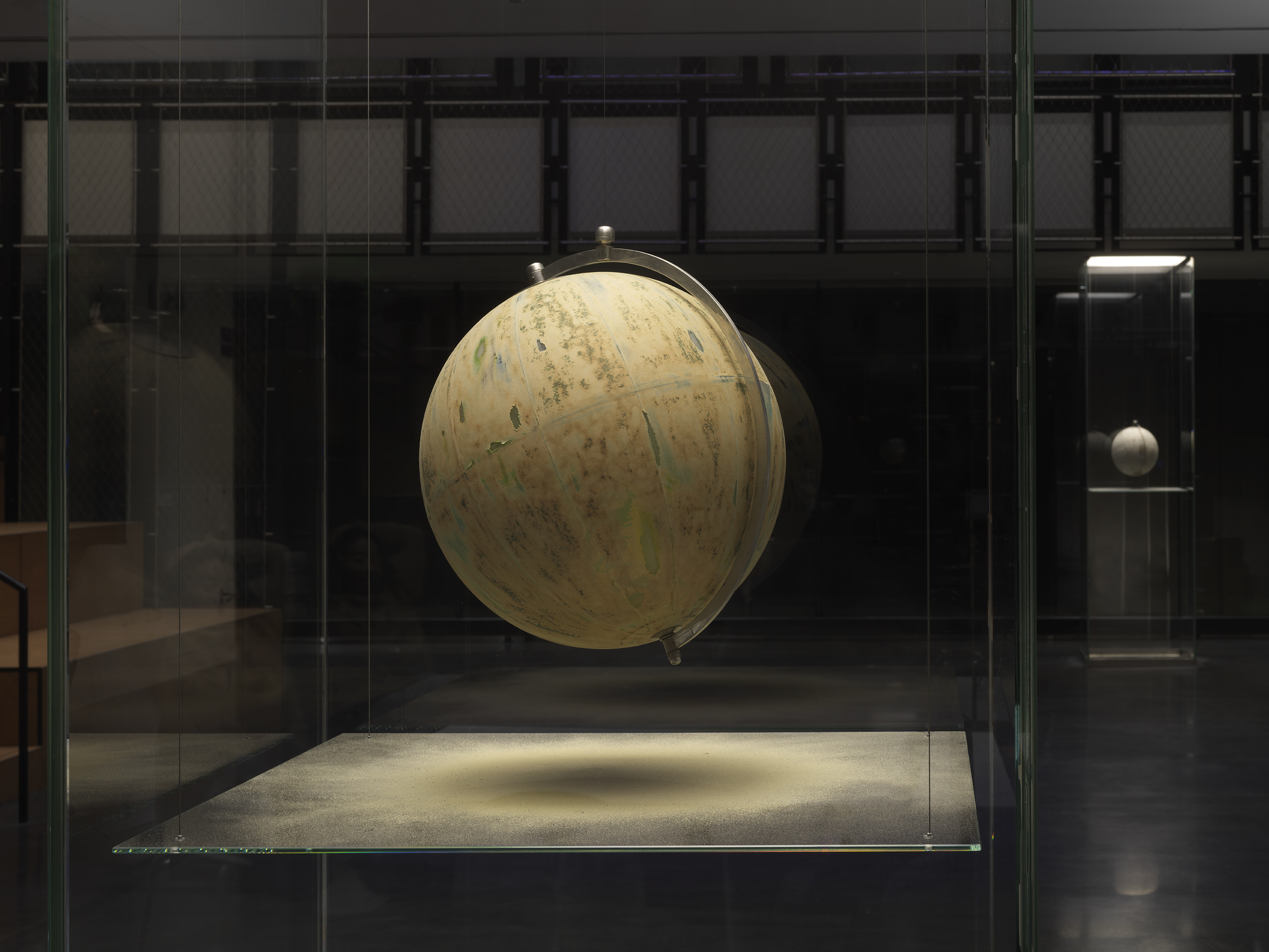 One painted globe suspended in a glass rectangular column and hangs over a flat surface that cuts the cylinder horizontally. Splatters of paint can be seen on top of the panel underneath the globe.
