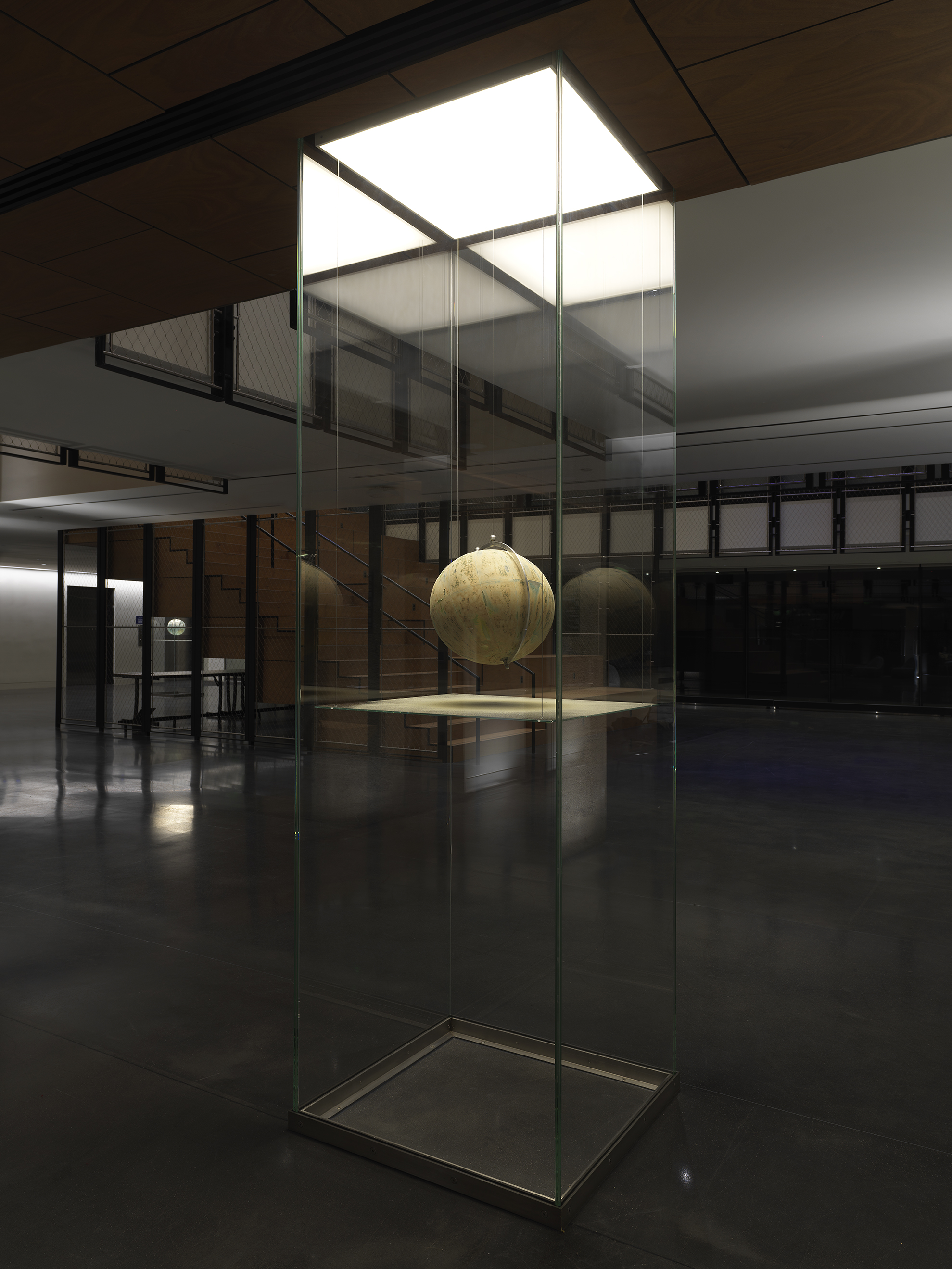 One painted globe suspended in a glass rectangular column and hangs over a flat surface that cuts the cylinder horizontally. Splatters of paint can be seen on top of the panel underneath the globe.