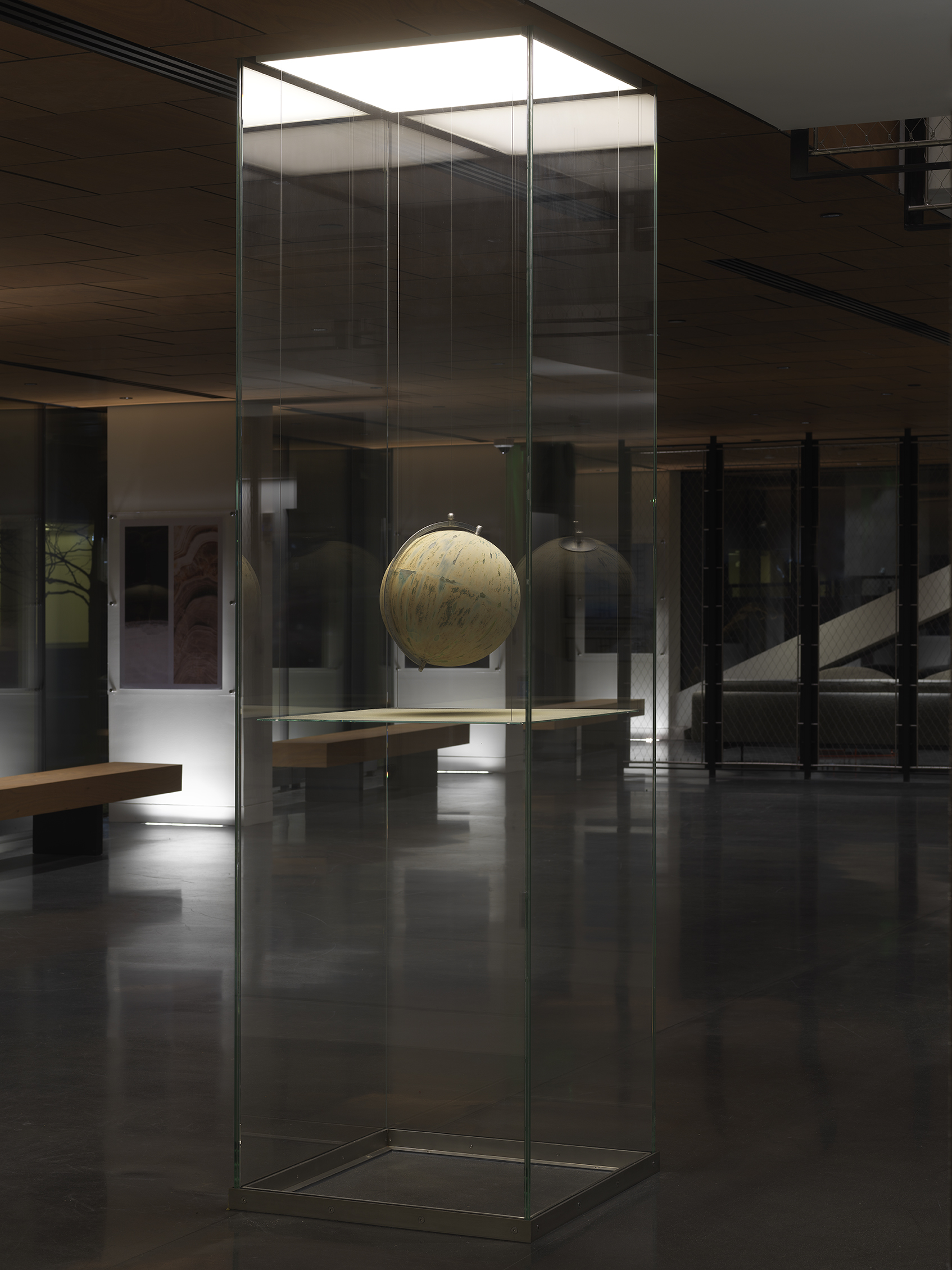 One painted globe suspended in a glass rectangular column and hangs over a flat surface that cuts the cylinder horizontally. Splatters of paint can be seen on top of the panel underneath the globe.