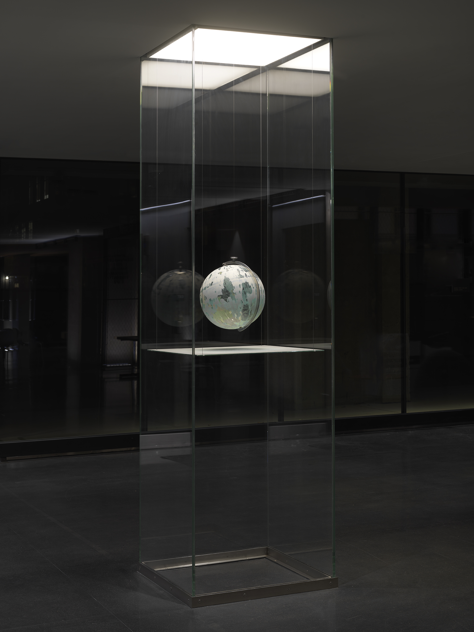 One painted globe suspended in a glass rectangular column and hangs over a flat surface that cuts the cylinder horizontally. Splatters of paint can be seen on top of the panel underneath the globe.