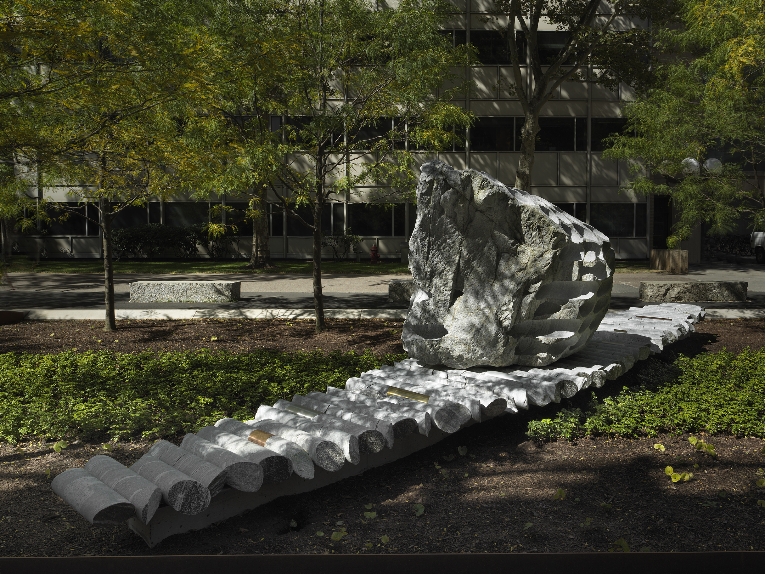 A large rock sculpture rests on along strip of stone cylinders in a green space. The rock has a series of holes removed from its core, with the cylinders that were removed making up the strip it rests on. Shadows and speckles of light are cast on the sculpture with a pathway, trees, and a gray building in the background.